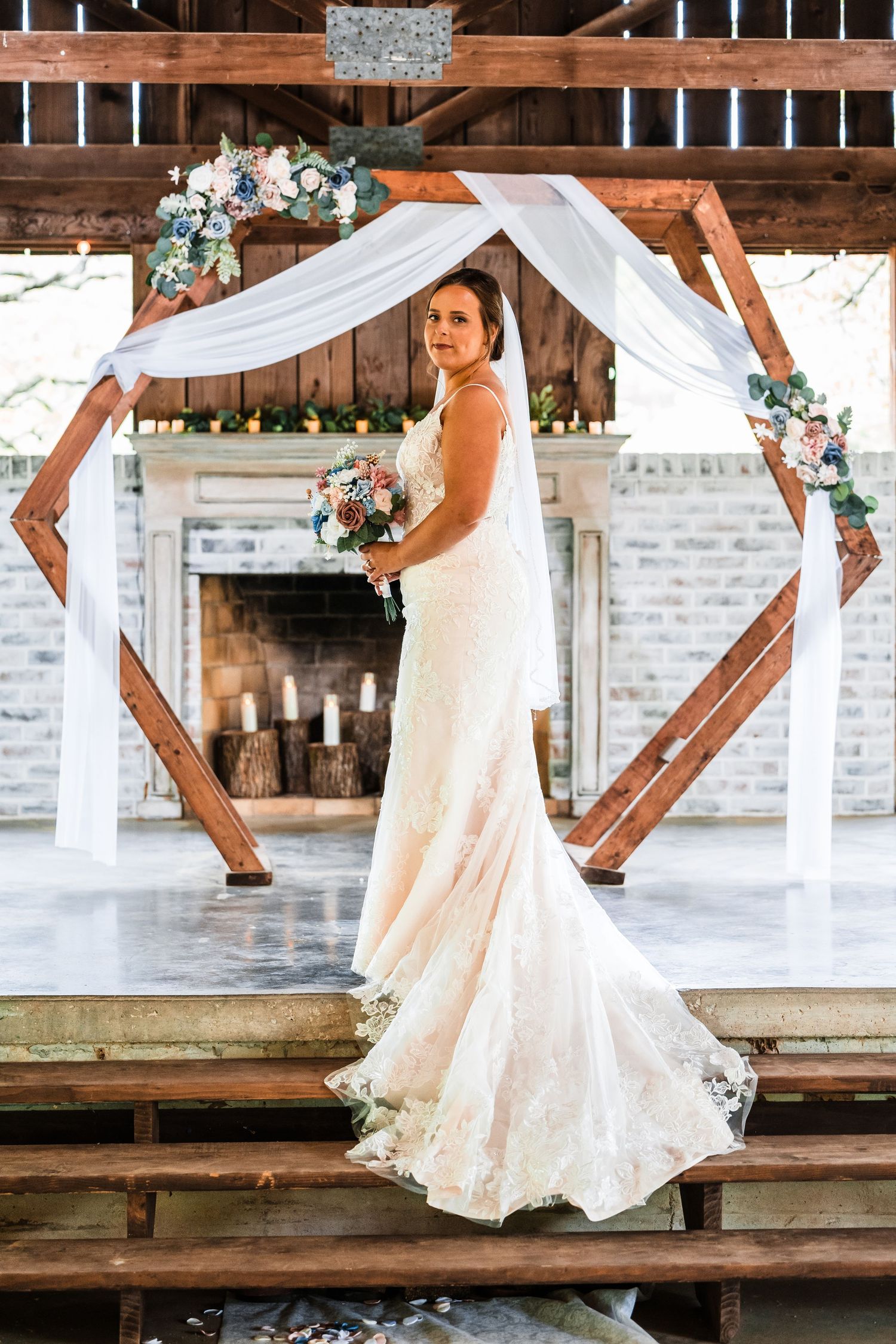 Elegant wedding ceremony setup with hexagonal wooden arch draped in white fabric and floral arrangements in rustic venue.
