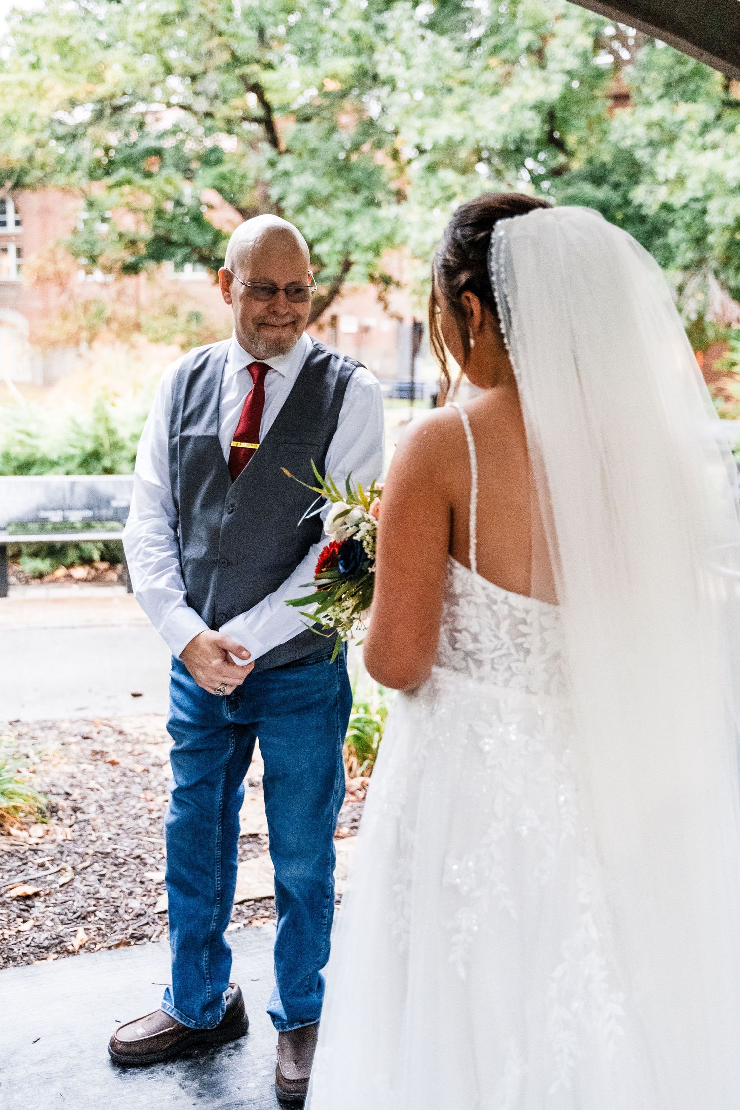 A touching first look moment between a bride in a white lace gown and a person wearing jeans and a vest.