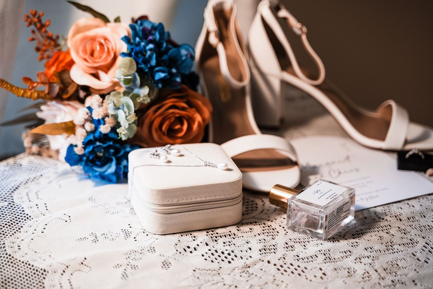 Wedding accessories arranged on lace tablecloth featuring blue and peach floral details.