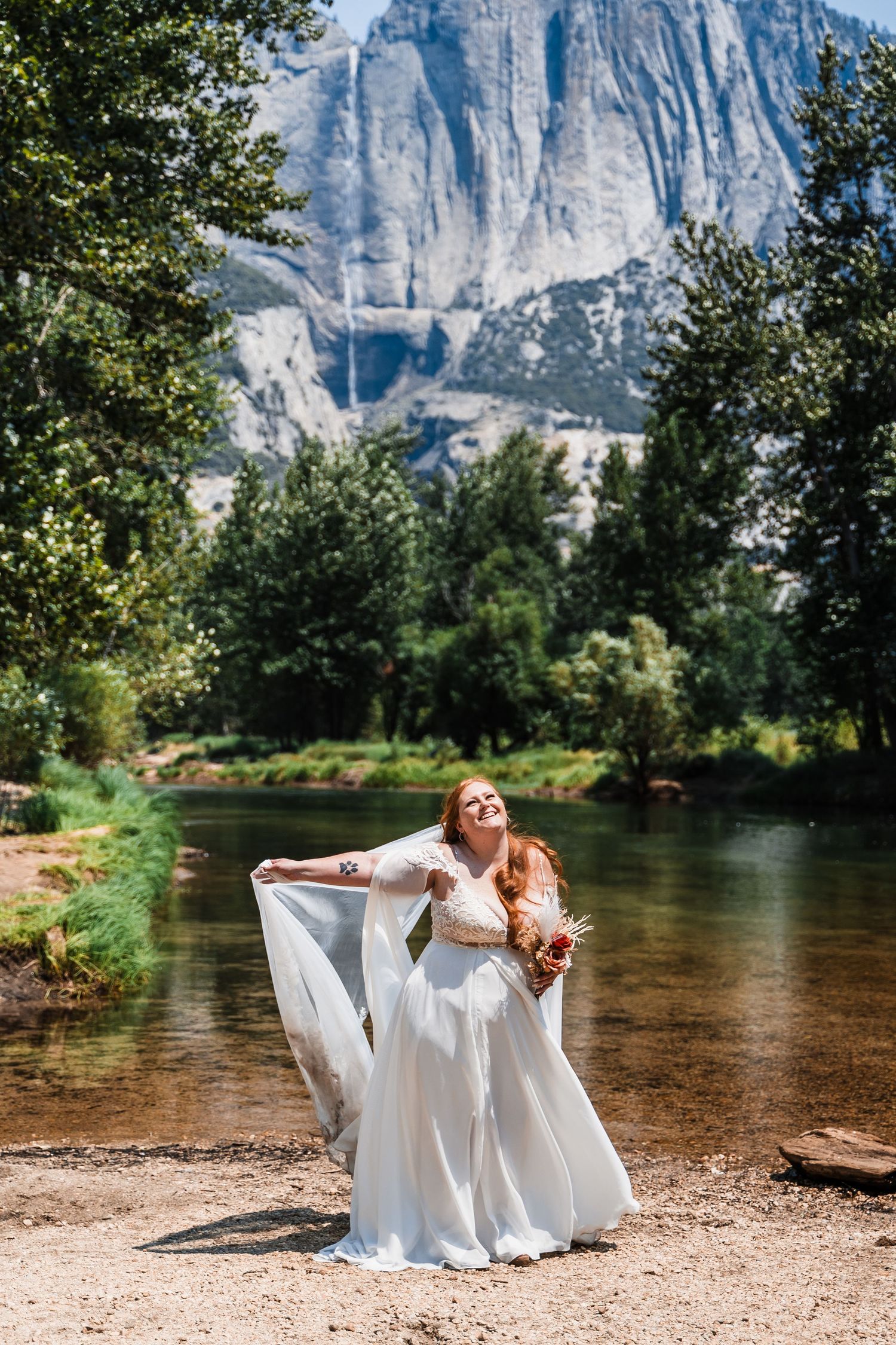 Bride in flowing white dress poses playfully by river with majestic Yosemite cliffs in background.