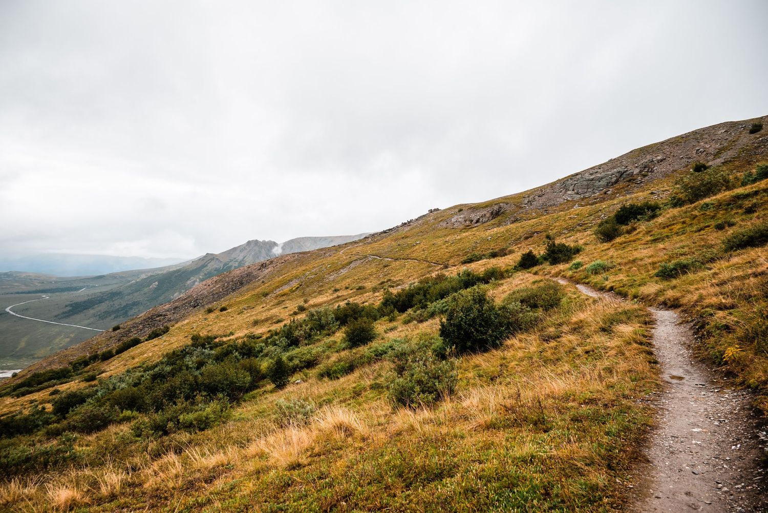 A winding dirt trail leads through golden mountain slopes under cloudy skies in a rugged highland landscape.