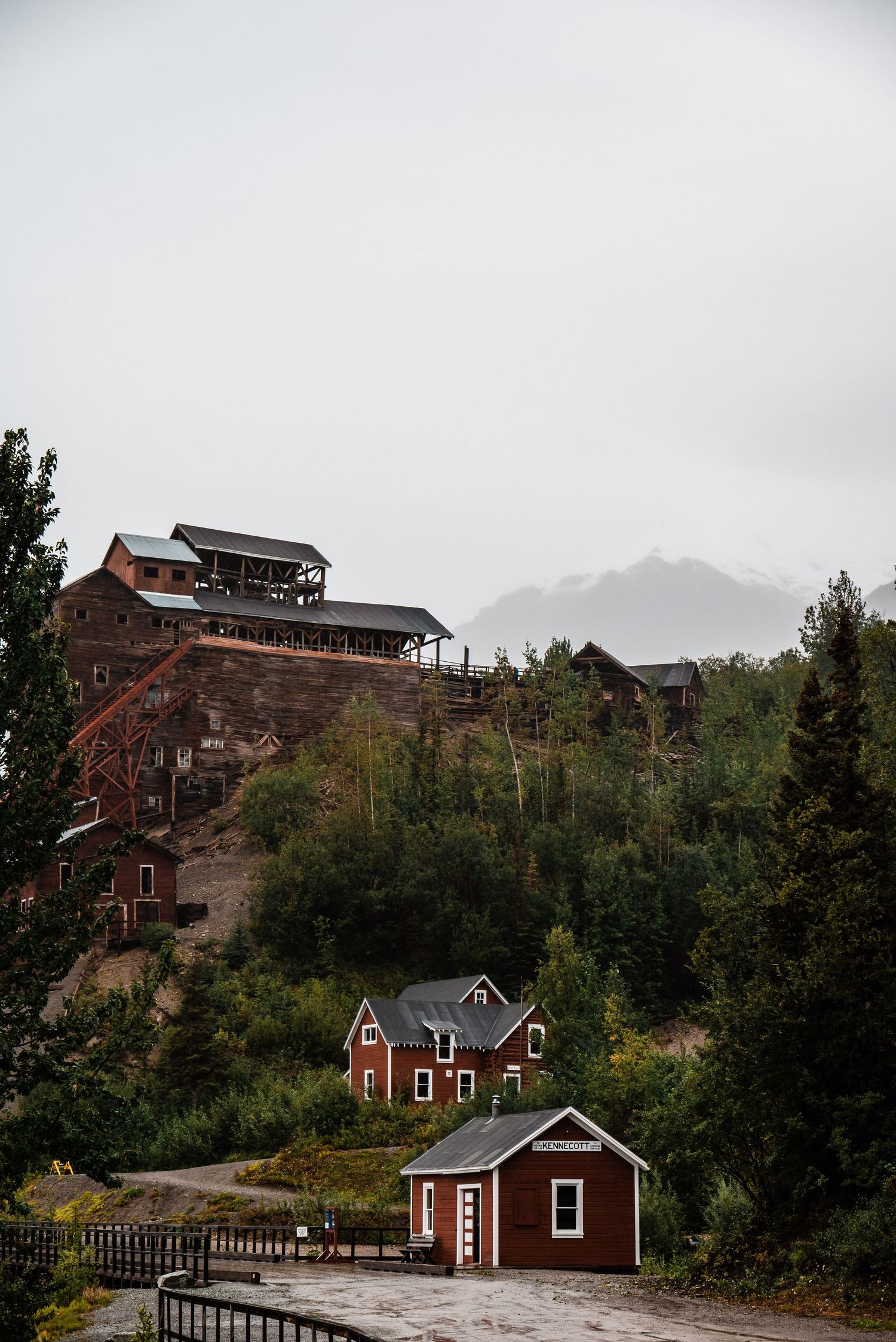 Historic mining buildings and red cabins perch on a forested hillside beneath misty mountain peaks.