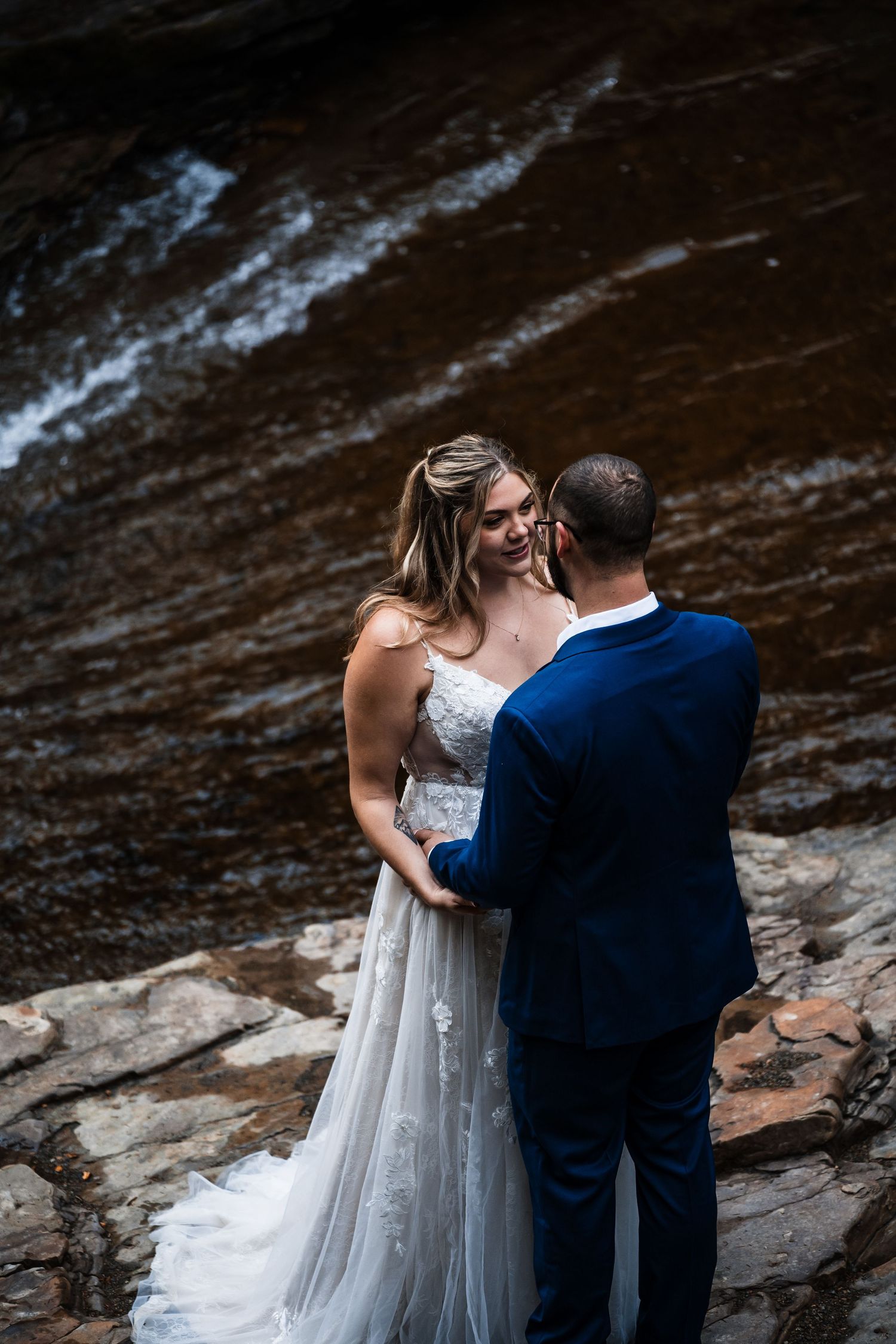 A couple in wedding attire stands together near a rocky riverbank in a romantic moment.