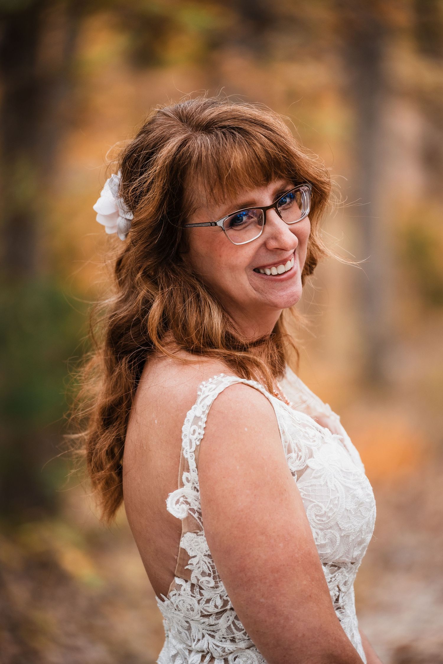A white lace wedding dress shown in a natural outdoor setting with autumn colors in the background.