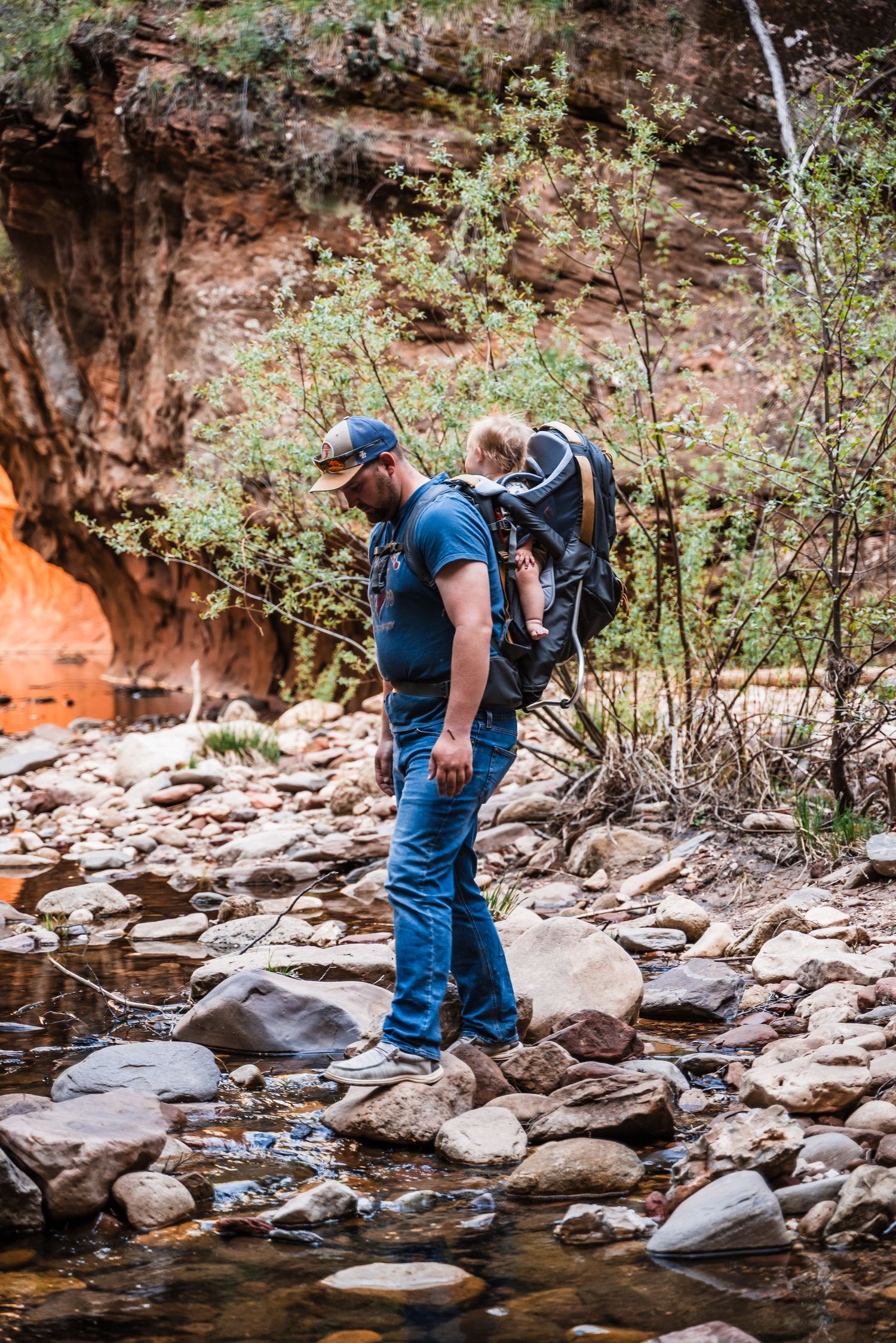 A hiker navigates rocky terrain along a river bank near a red sandstone cliff while carrying a child carrier backpack.