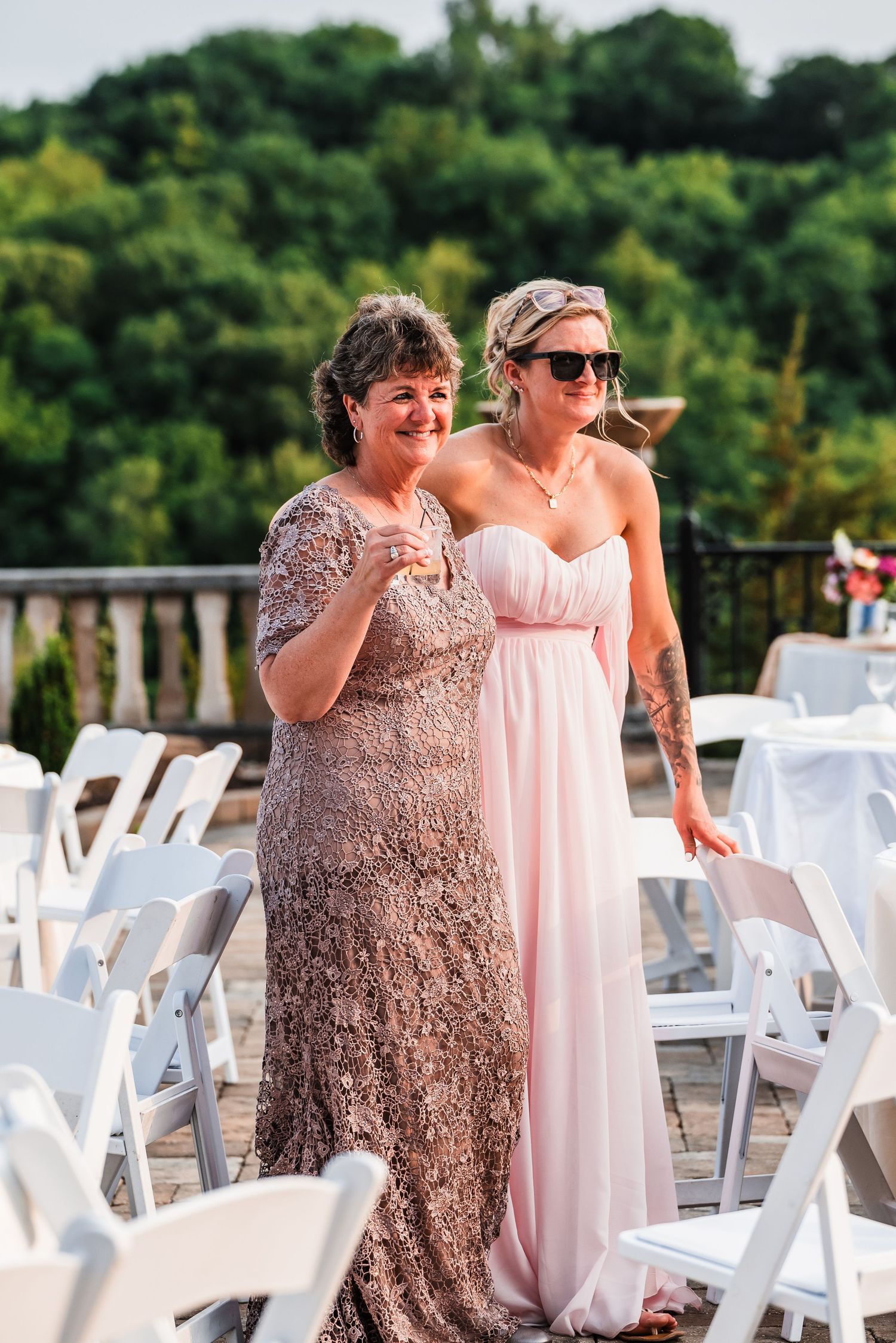 Two wedding guests in formal dresses standing among white chairs on outdoor patio.