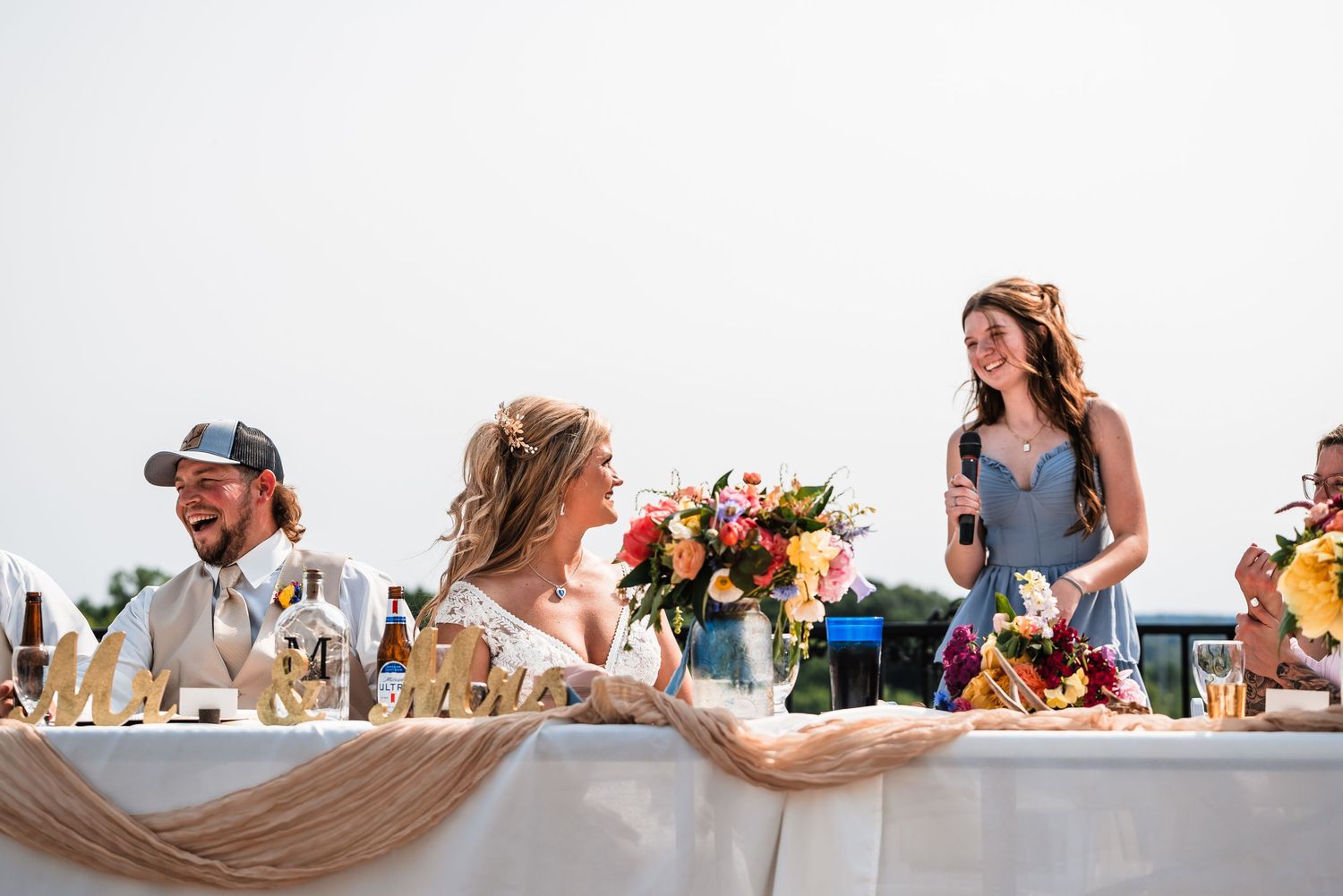 Wedding reception guests seated at a long table decorated with floral arrangements and tablecloths outdoors.