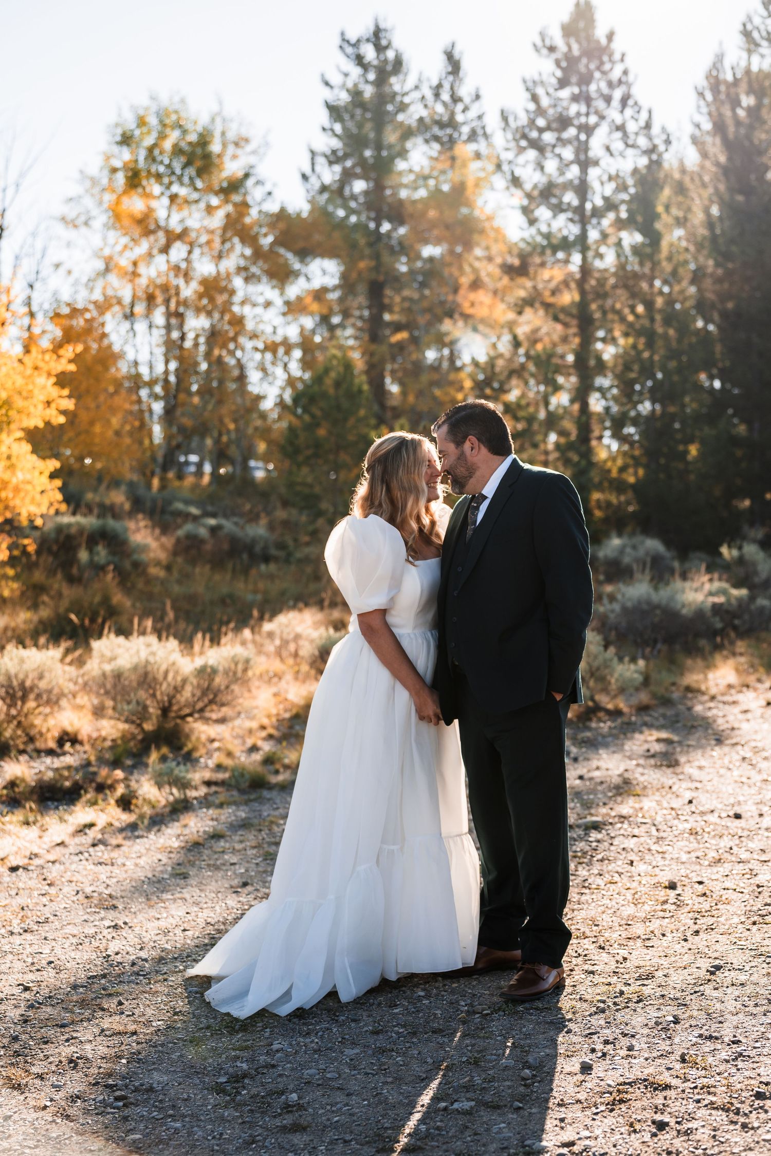 A couple in wedding attire sits together on a hillside path during golden hour sunset in a forest setting.