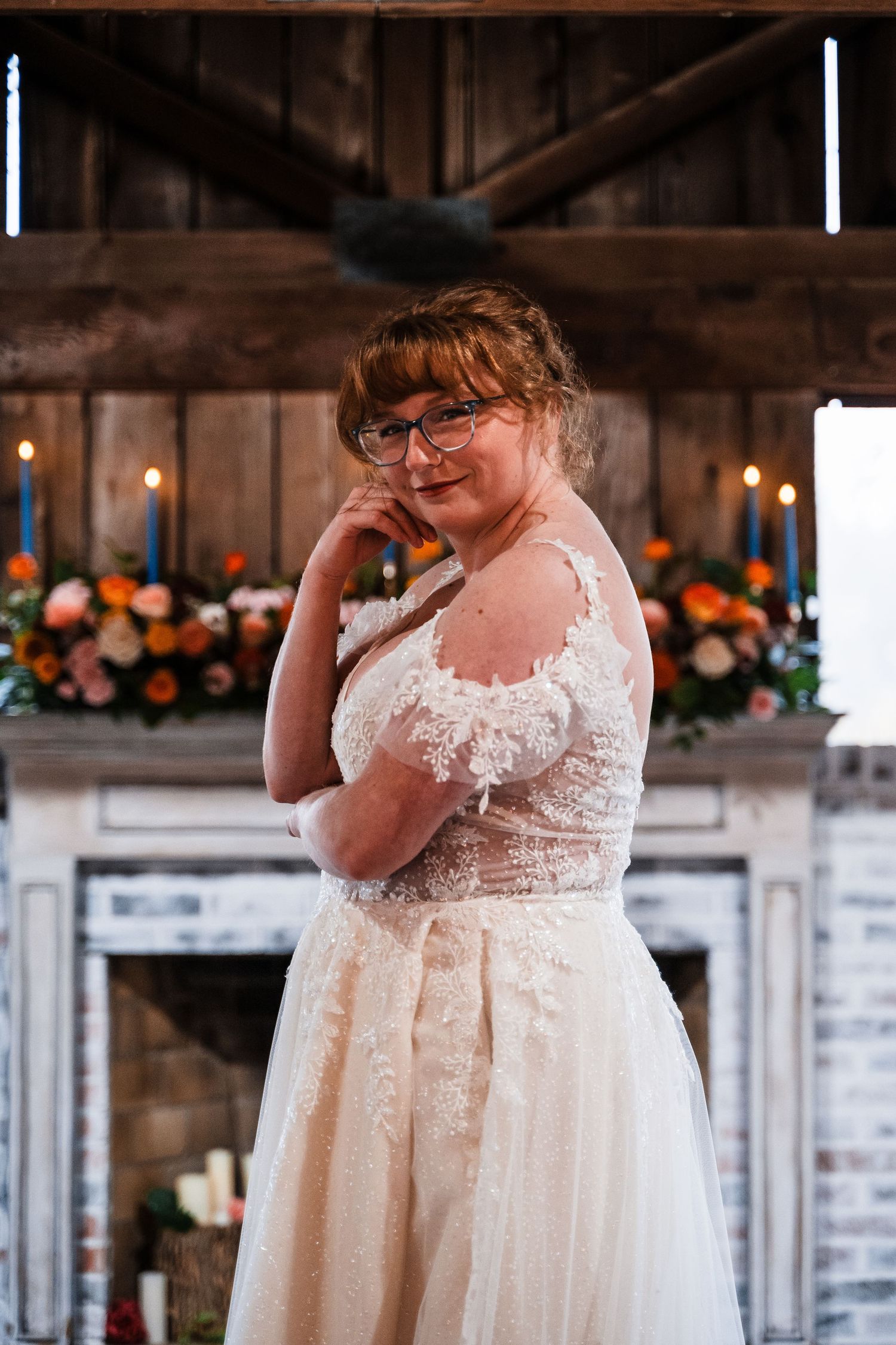 Bride in elegant lace wedding dress poses inside rustic barn venue with floral decorations.