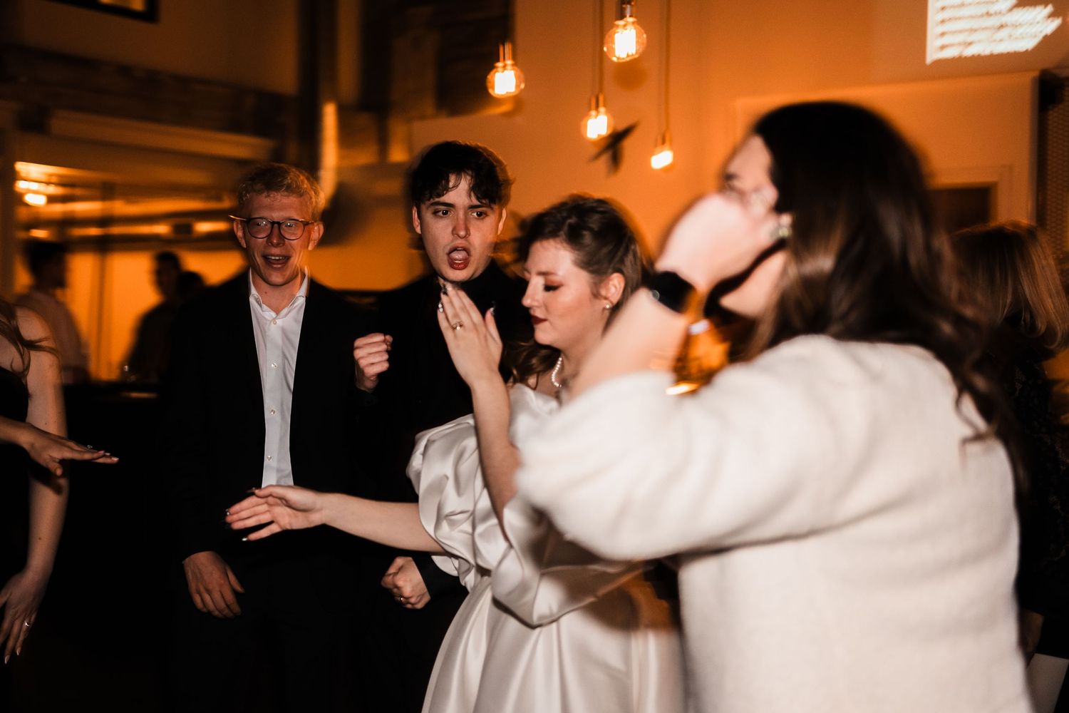 Wedding guests dancing and singing with joyful expressions at an indoor reception venue.