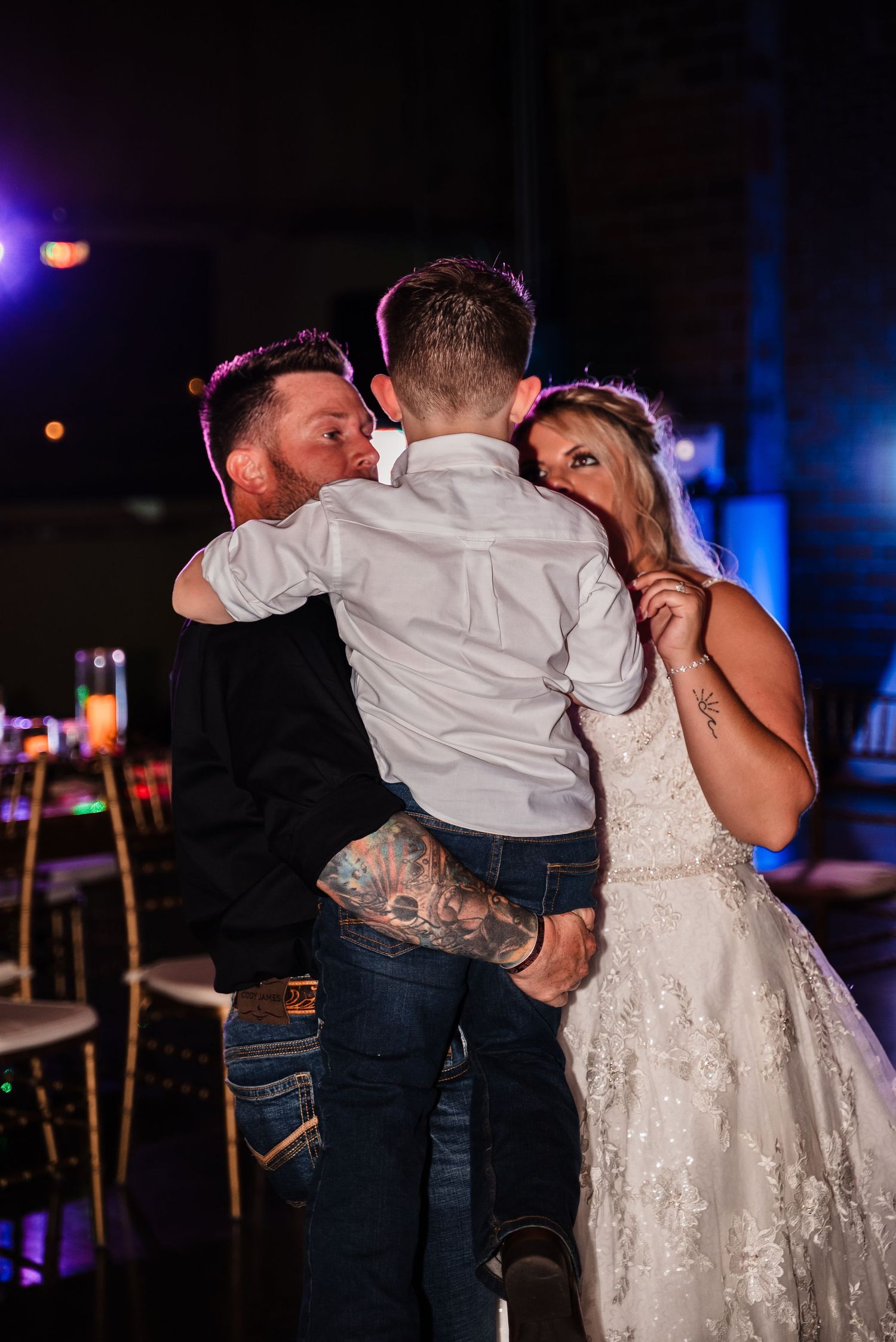 A sweet moment shared between couple in wedding attire and young child during an indoor reception with blue lighting.