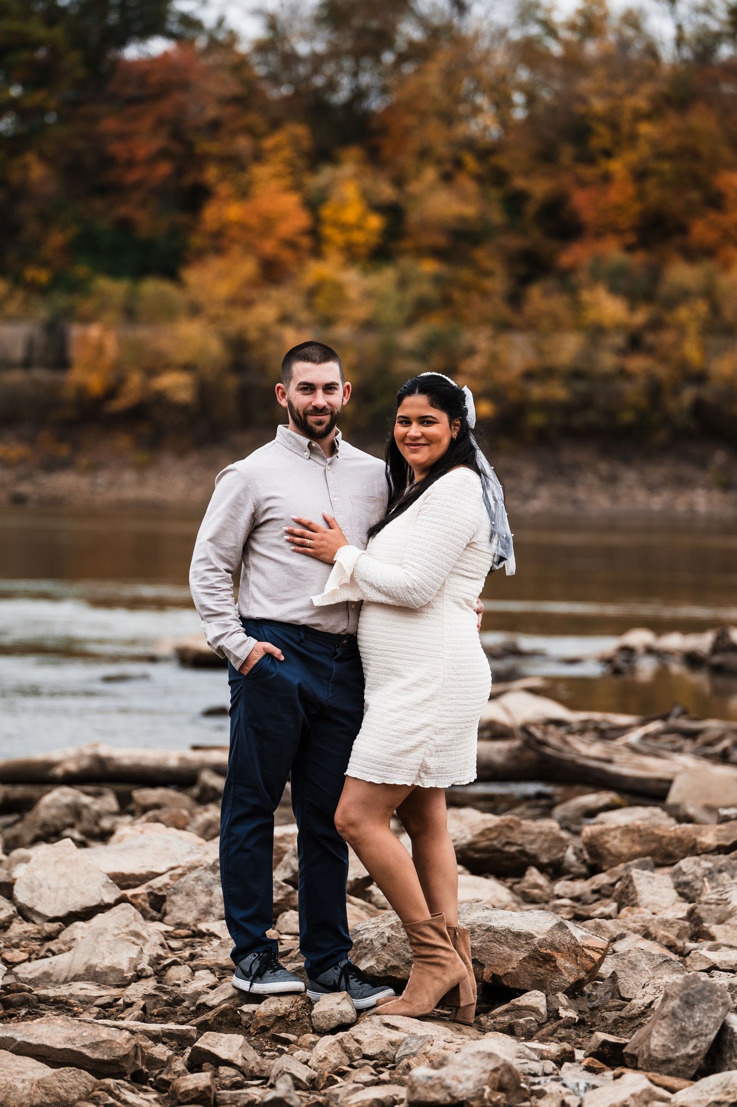 Couple standing on rocky shore during autumn with colorful foliage in background.