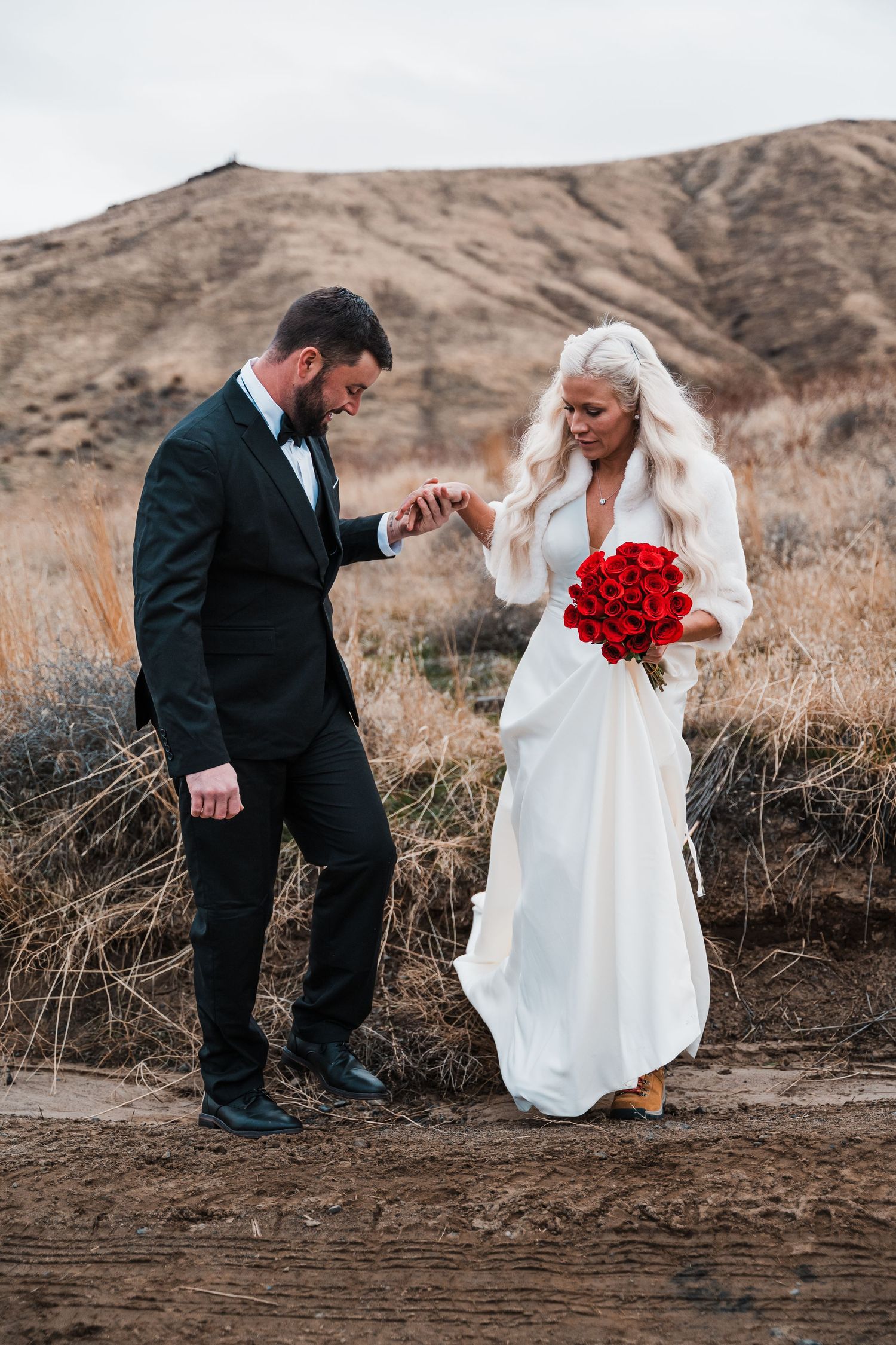 A wedding couple stands against desert hills as the groom helps steady the bride holding red roses.