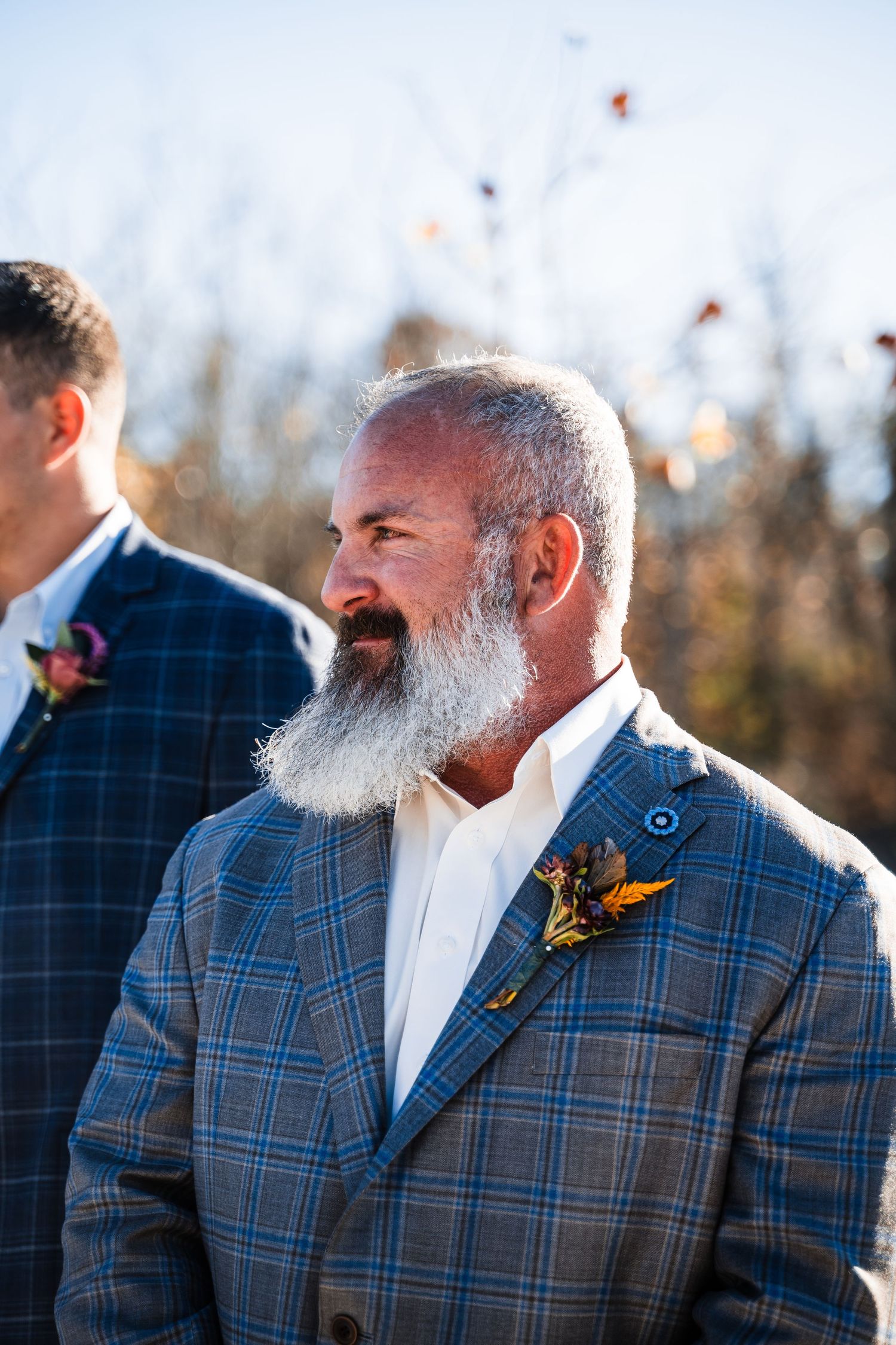 Person wearing plaid suit jacket with boutonniere smiling during outdoor winter ceremony.