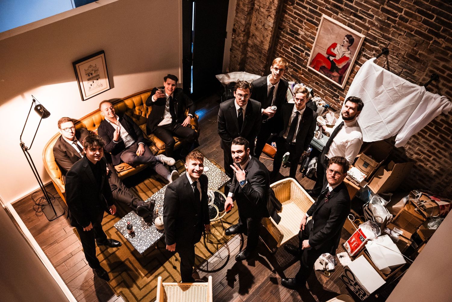 Wedding party in dark suits pose together in a rustic brick-walled room from above.