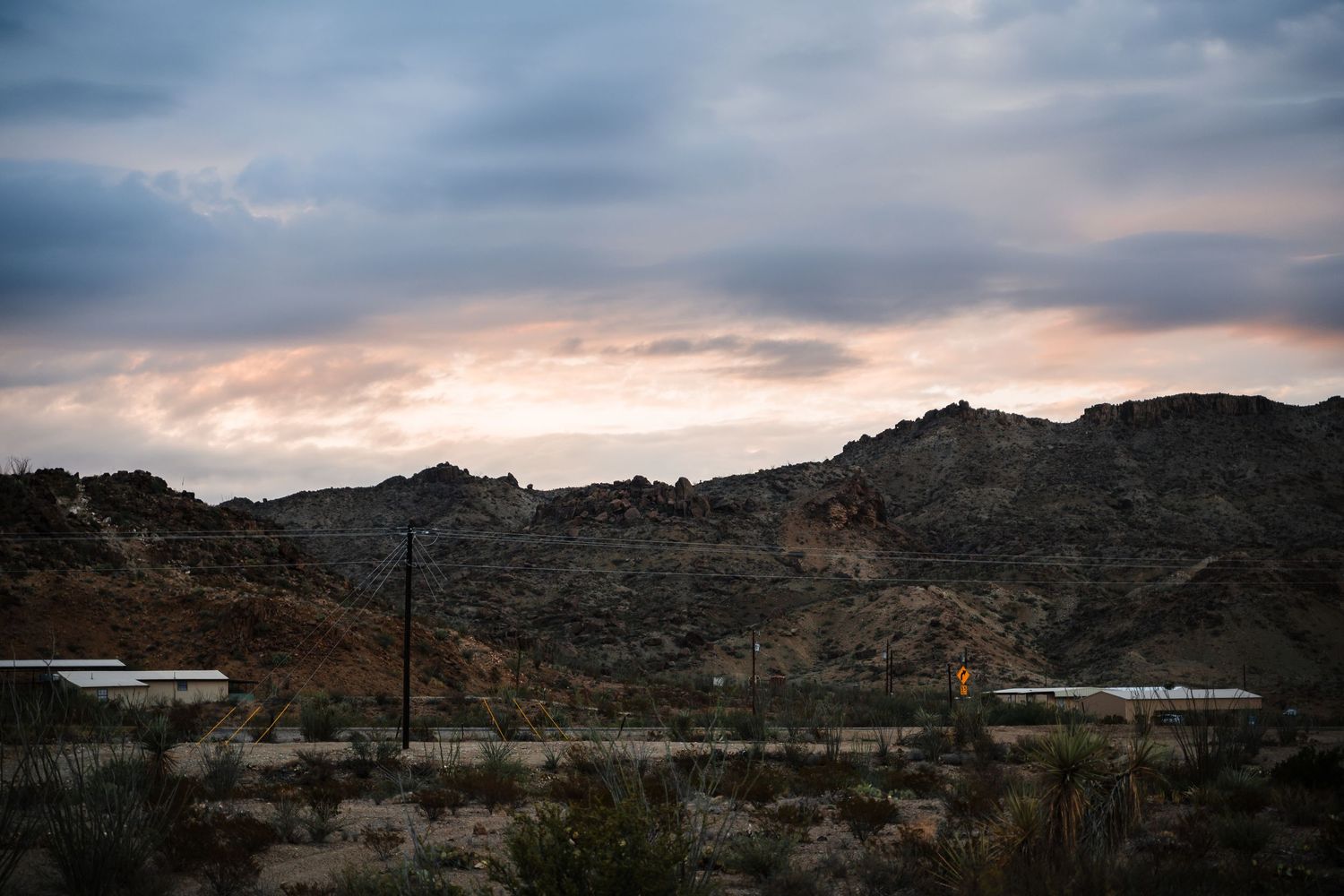 Desert mountain landscape with power lines silhouetted against a dramatic sunset sky with clouds.