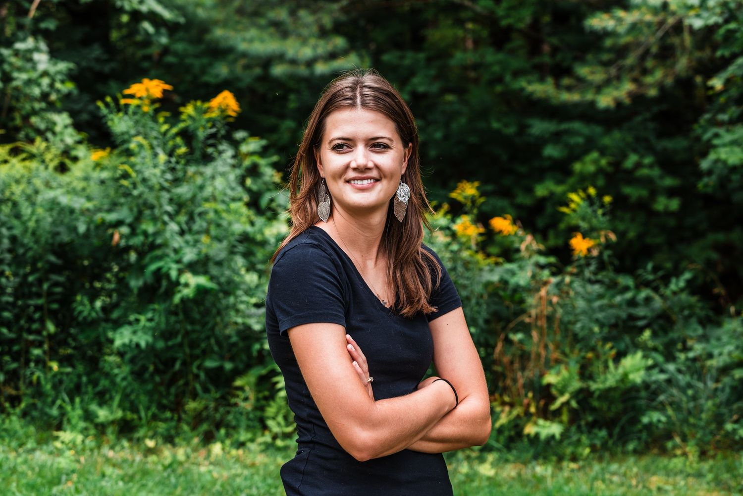 Someone in a black t-shirt poses with arms crossed in a garden with yellow flowers and greenery in the background.