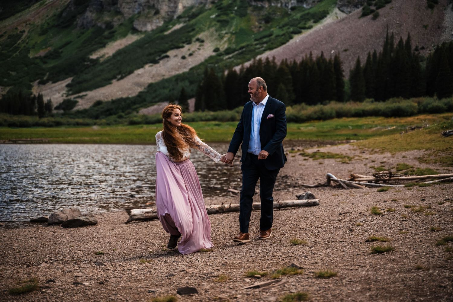 Couple holding hands by mountain lake during sunset with dramatic landscape behind them.