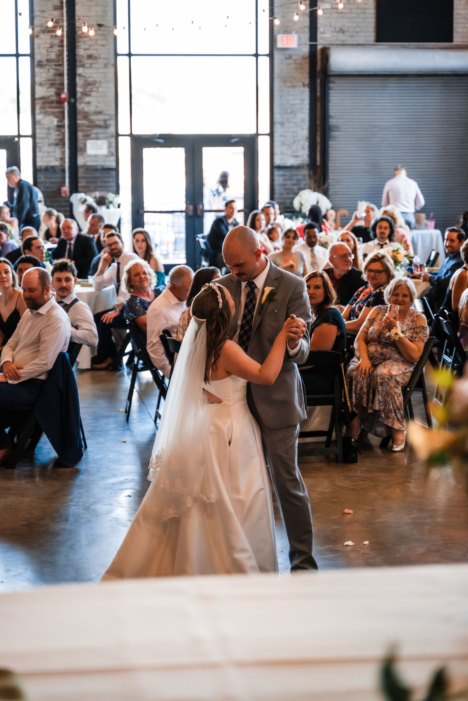 A couple shares their first dance at a wedding reception in an industrial venue while guests look on and applaud.