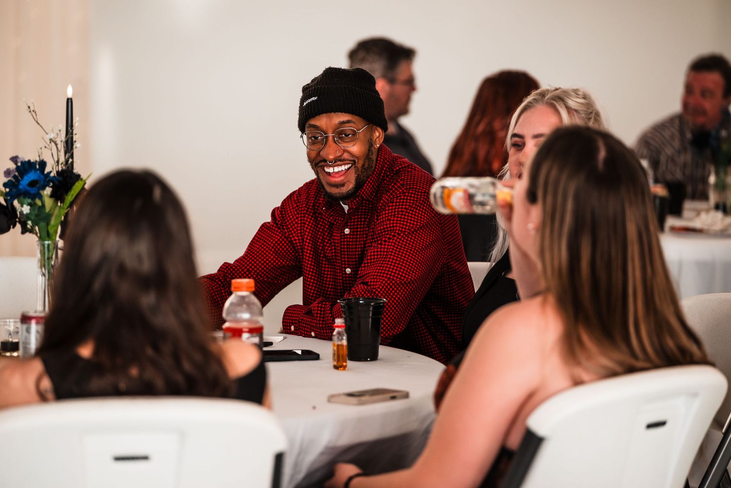 A smiling man in a red shirt and black beanie sits at a white table, laughing with a diverse group at an indoor gathering.