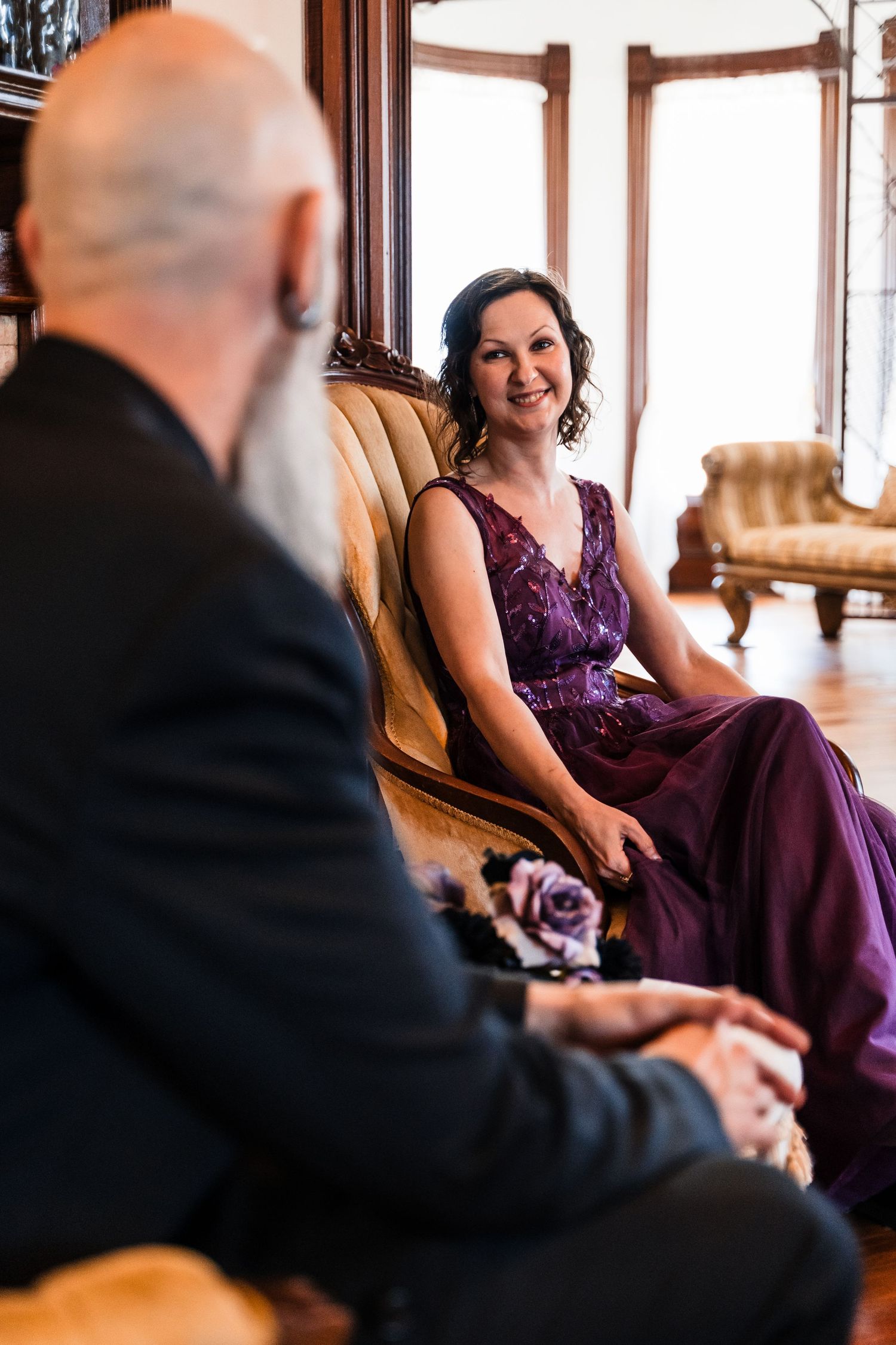 Bride in purple sequin gown smiling during intimate wedding ceremony seated on vintage sofa.