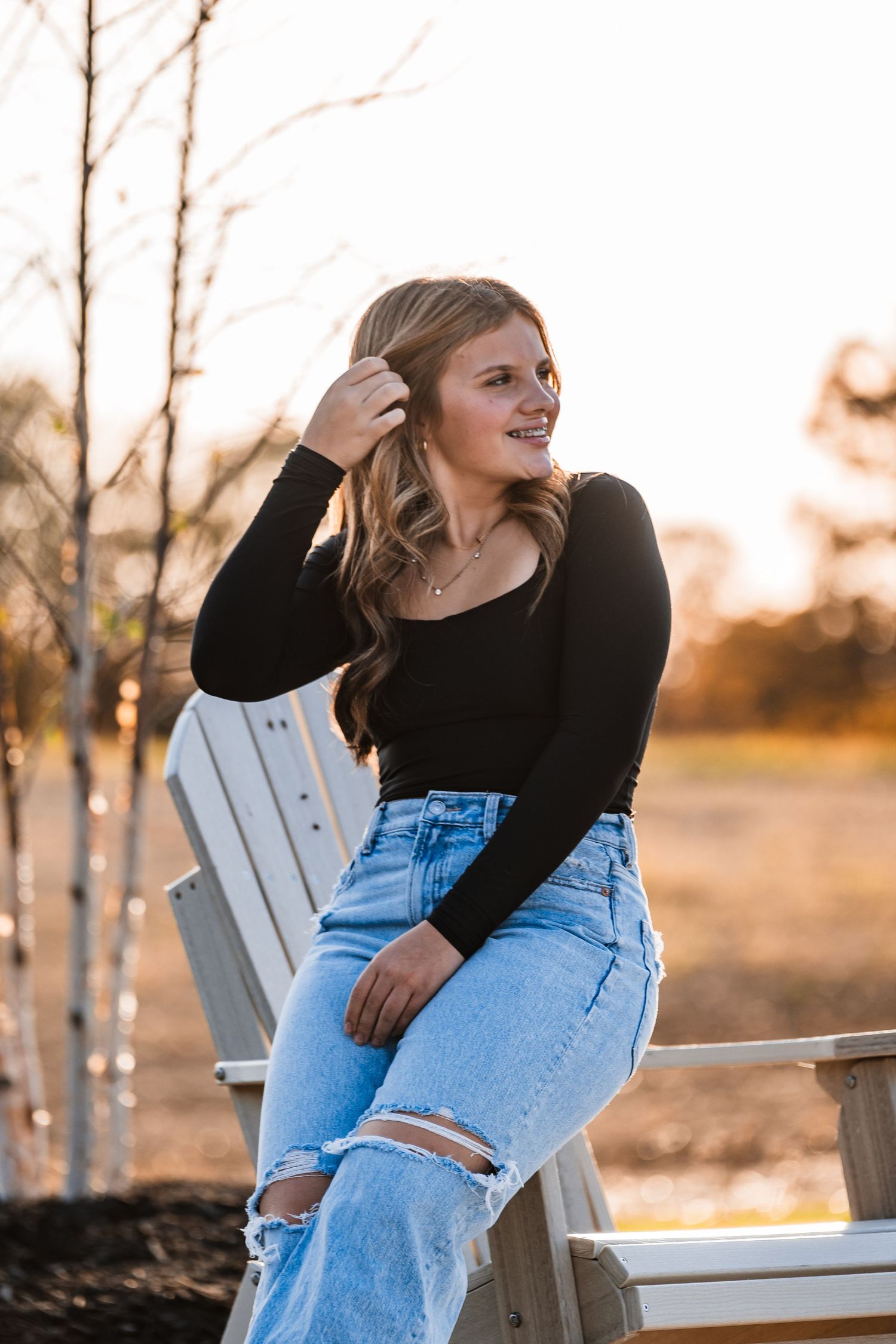 Person in ripped jeans and black sweater sits on white Adirondack chair during golden sunset.