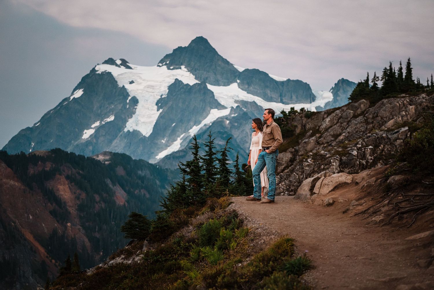A panoramic mountain landscape with snow-capped peaks and winding hiking trails through rugged alpine terrain.