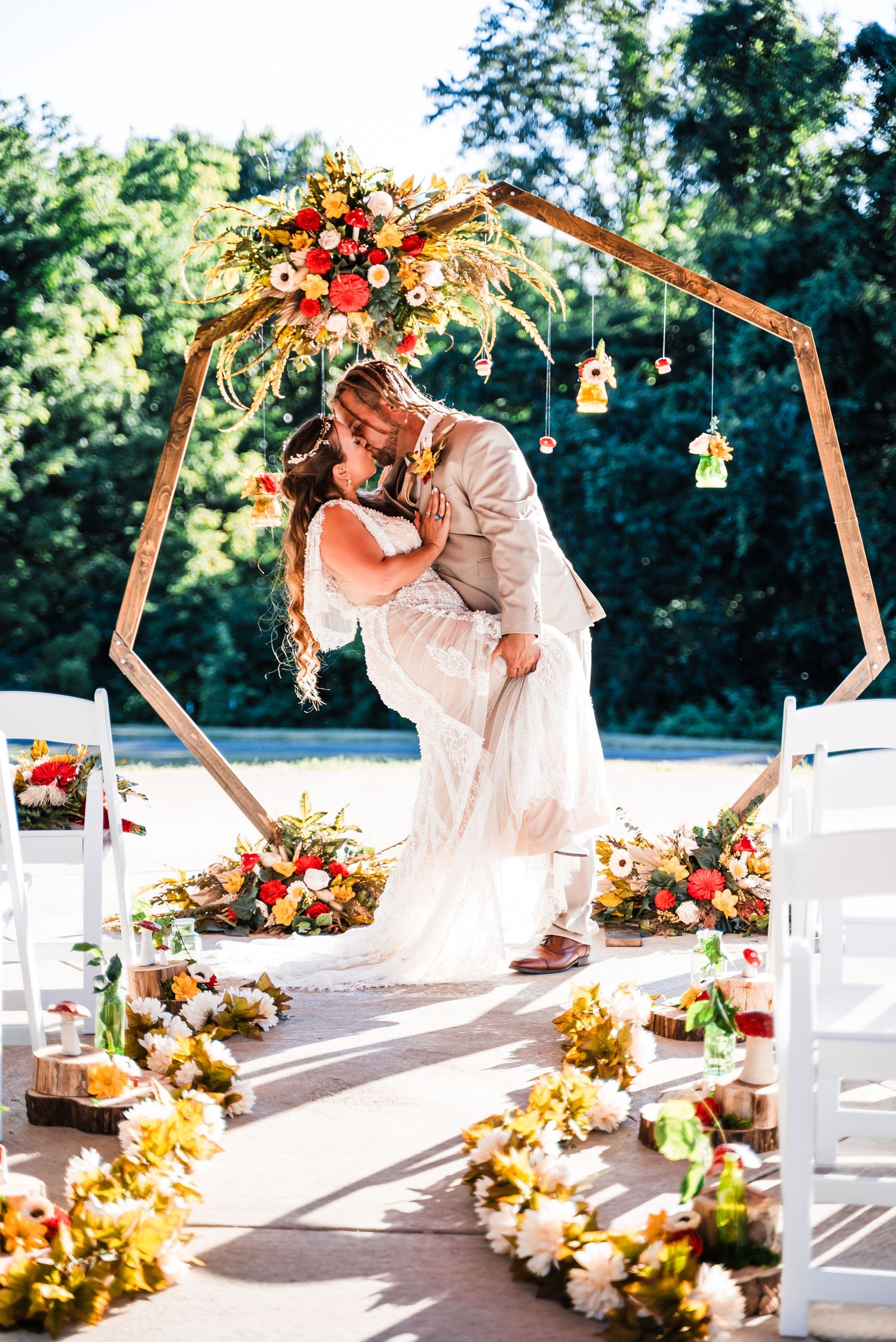A decorated outdoor wedding ceremony setup with wooden arch, white chairs, and autumn-colored floral arrangements.