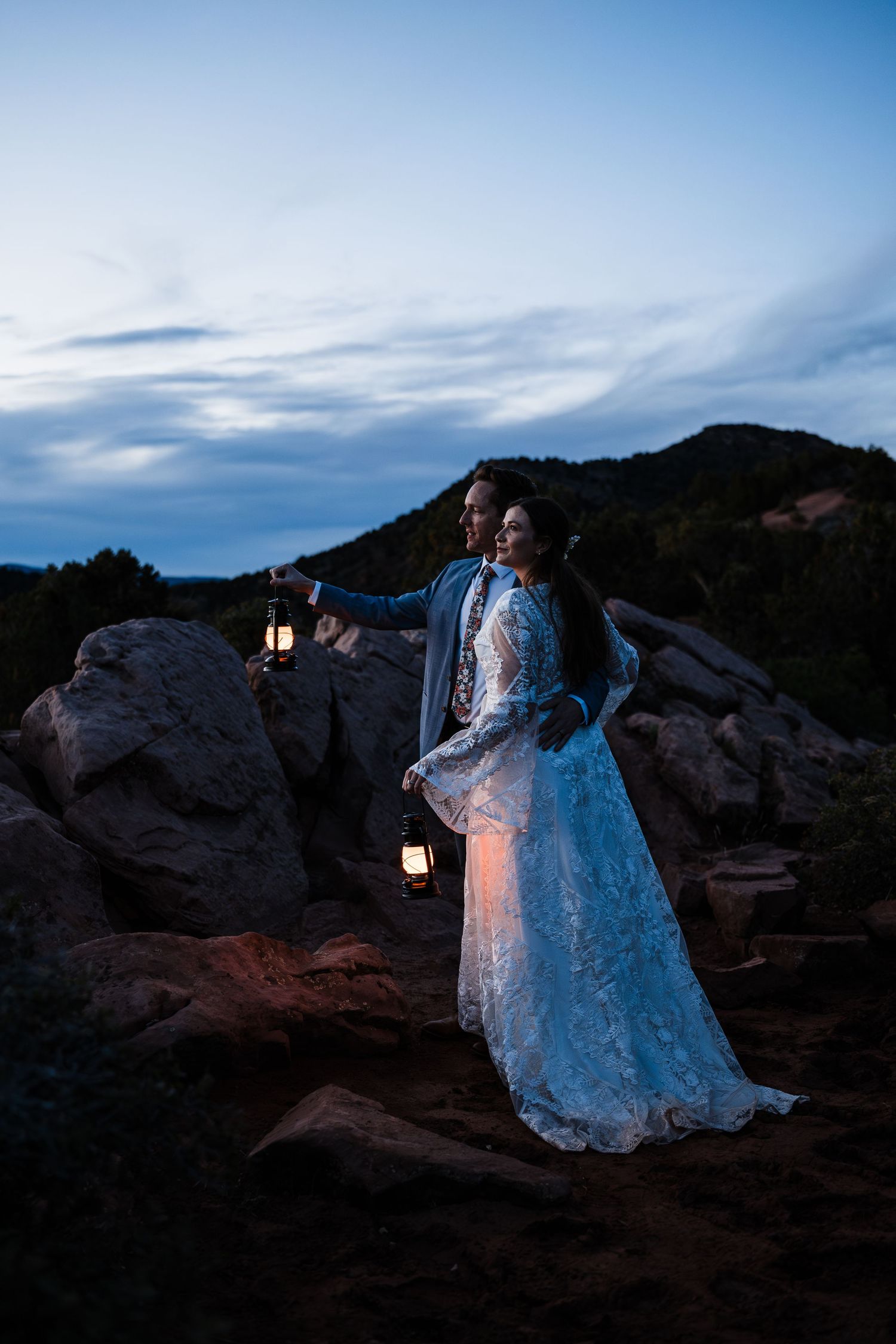 Bride and groom holding vintage lanterns on red rocks at night with glowing mountain backdrop.