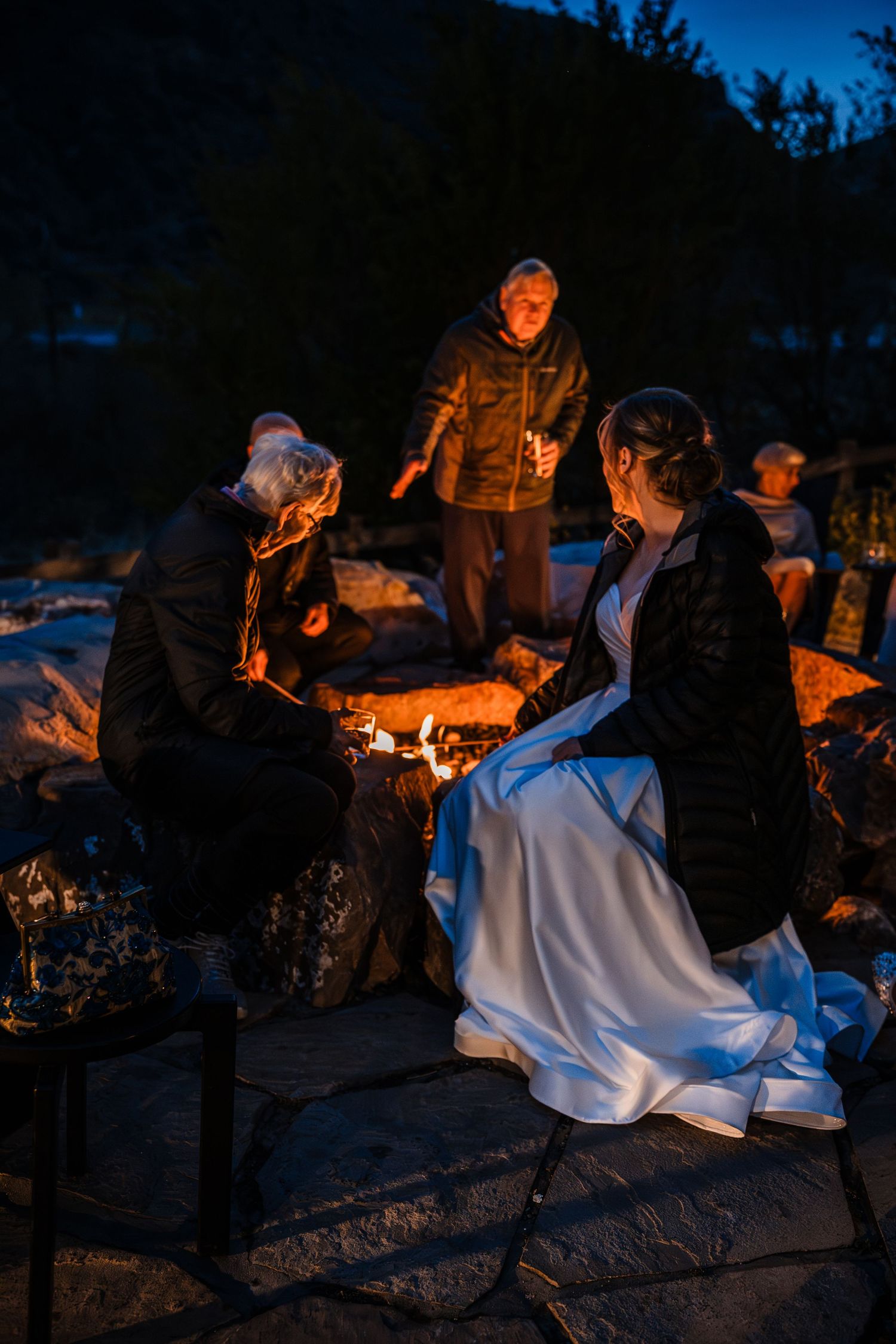 Bride in white gown sits by a glowing campfire with guests during a nighttime outdoor wedding.