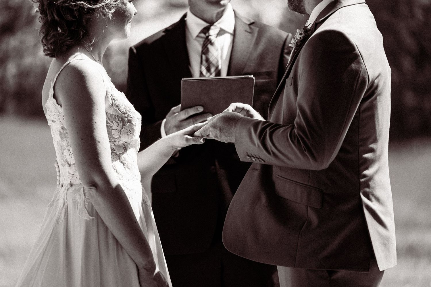 Black and white wedding ceremony photo of couple exchanging rings with officiant holding book in background.