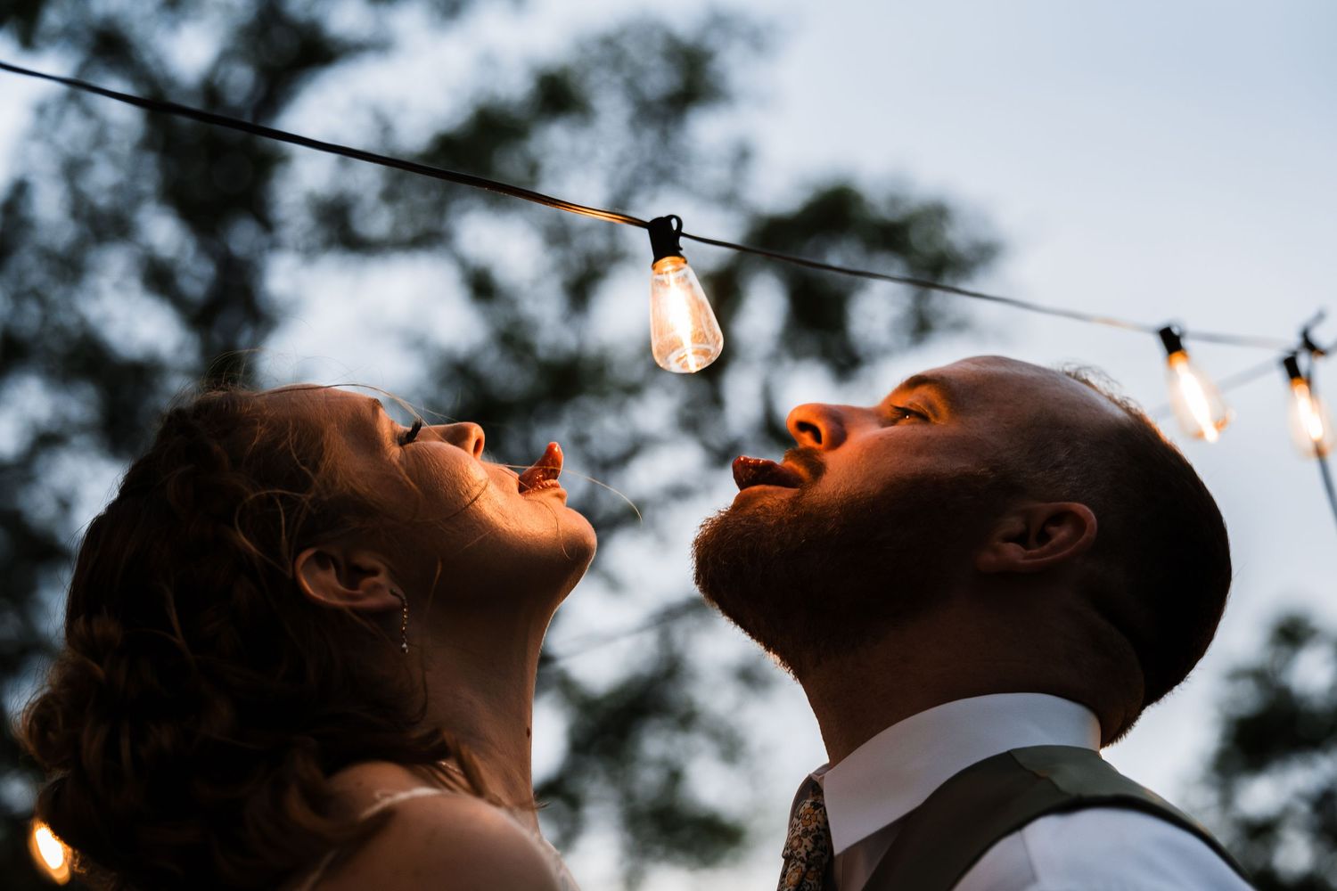 Bride and groom gazing up at string lights during an outdoor evening wedding celebration.