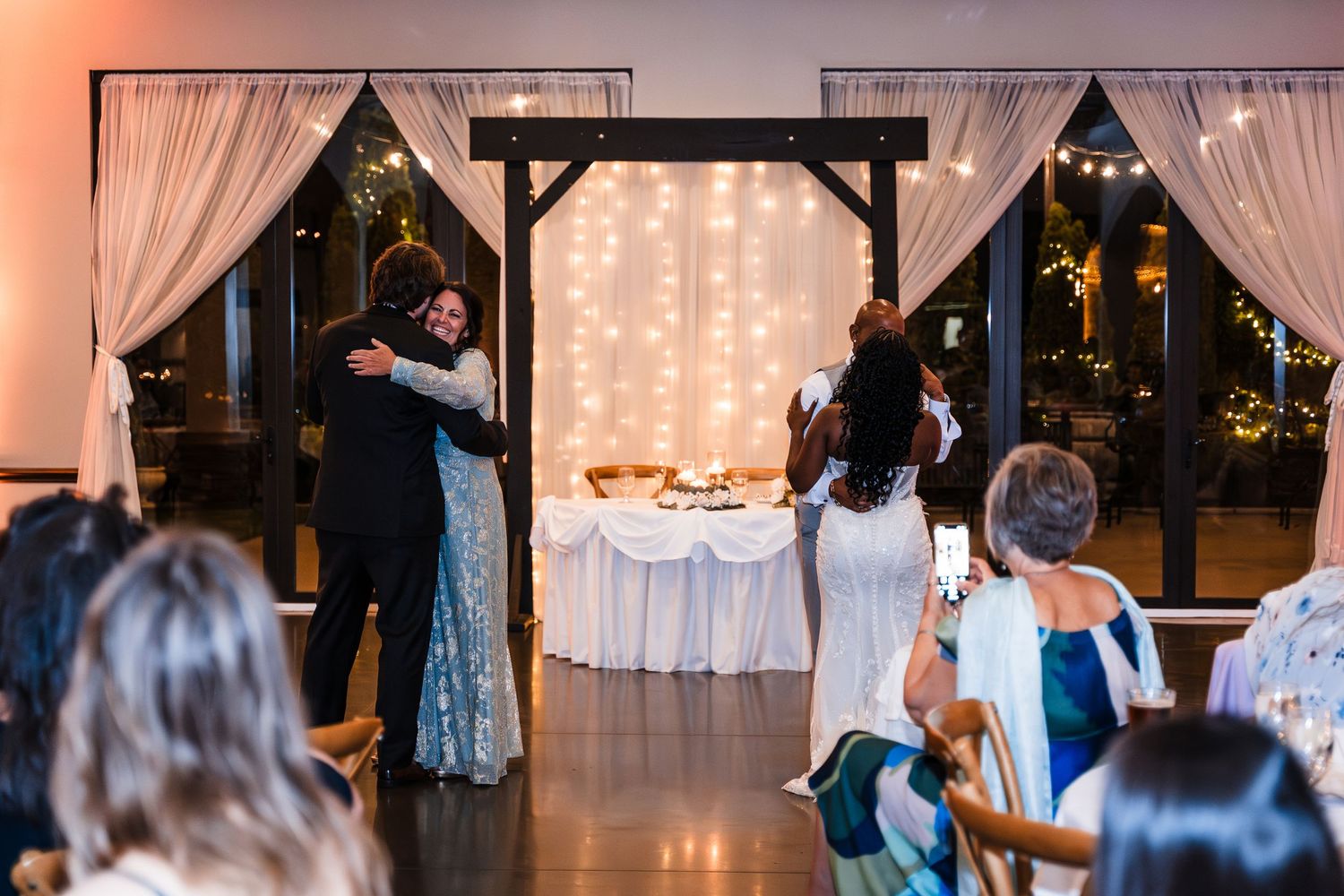 Couple shares warm embrace on dance floor as wedding guests watch and photograph the moment.