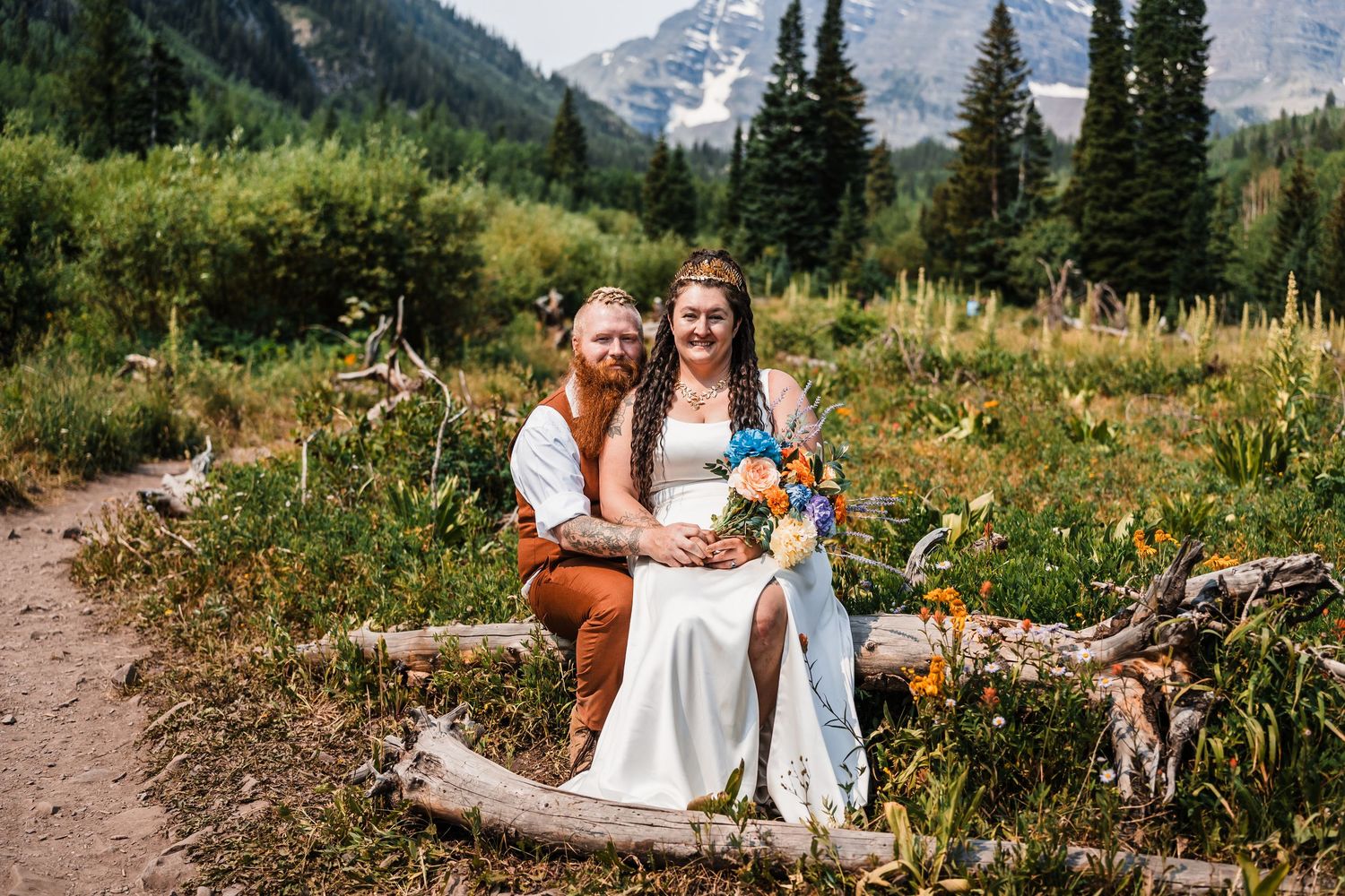 Eloped couple sits on fallen log surrounded by wildflowers and mountains in Colorado wilderness.