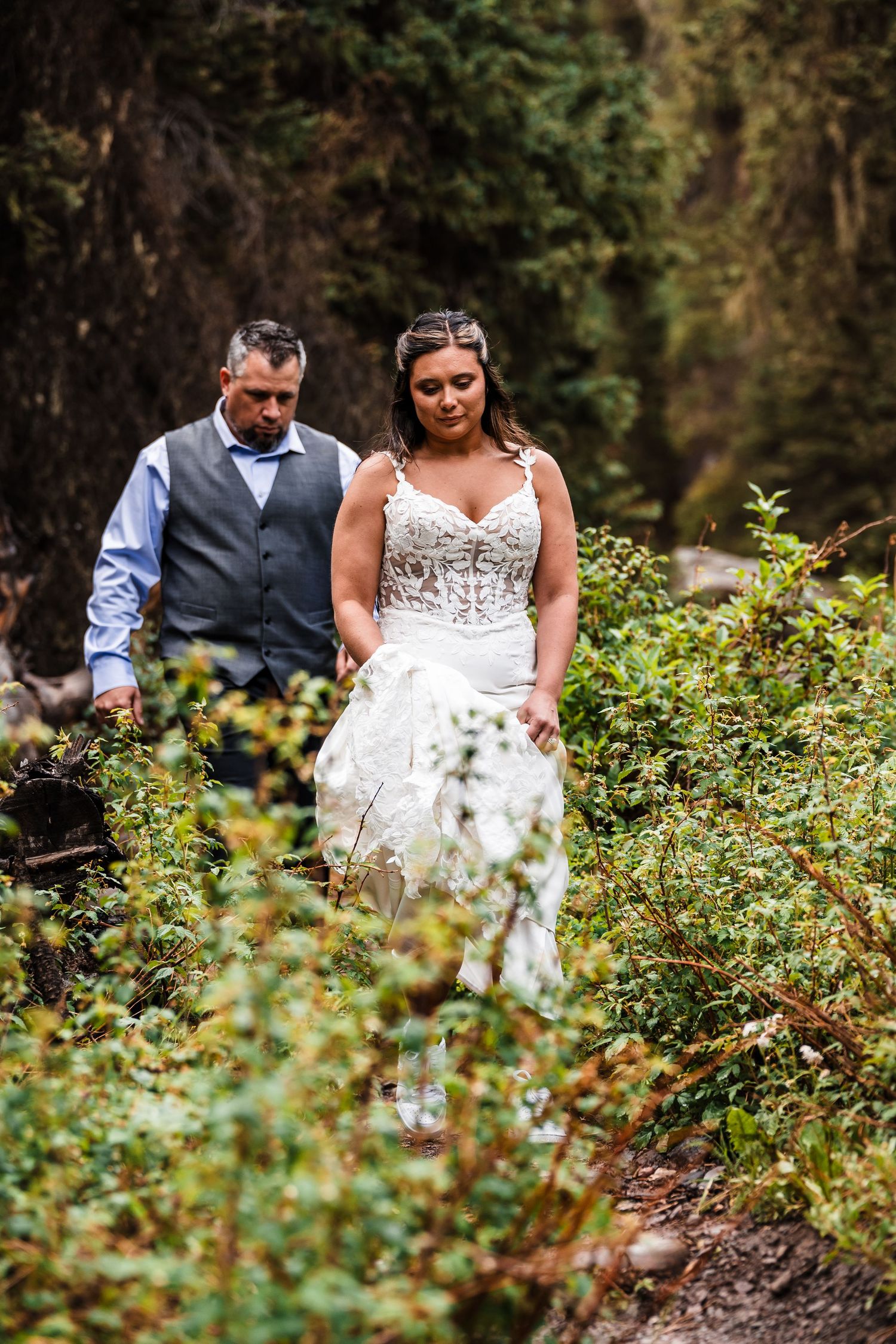 Bride in lace gown and groom walk hand in hand through lush green forest foliage.