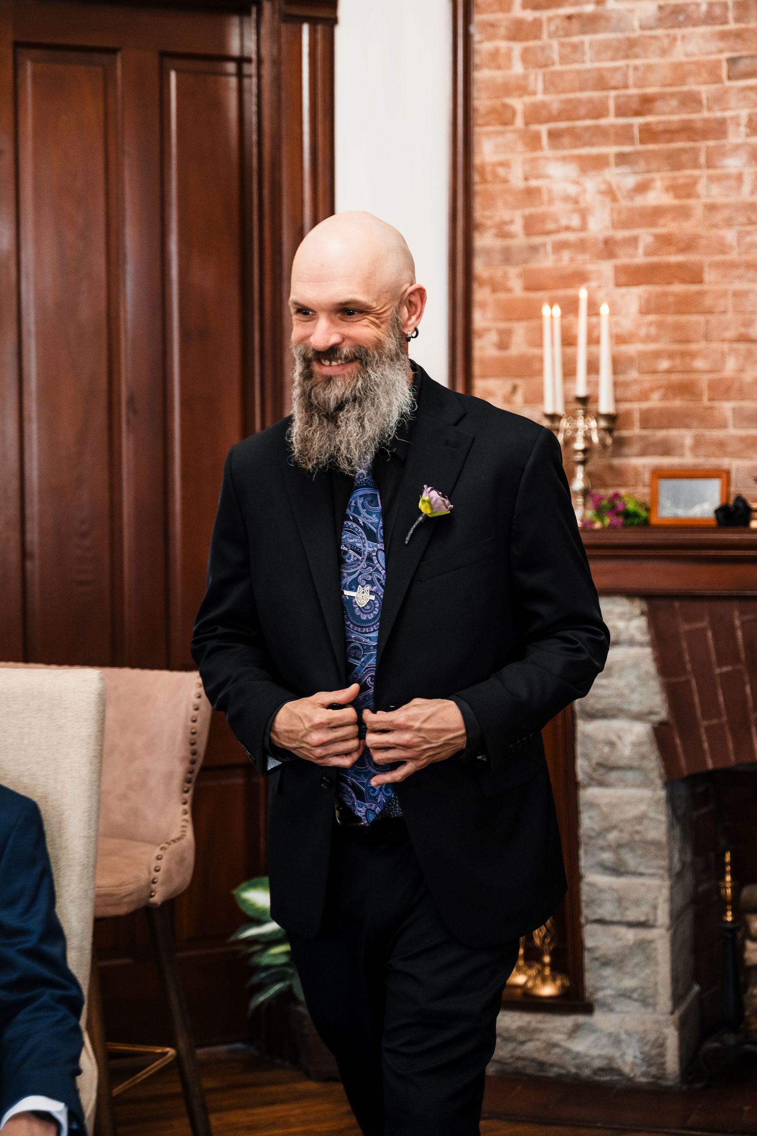 Smiling bald groom with beard wearing black suit and blue floral tie with boutonniere at wedding reception.