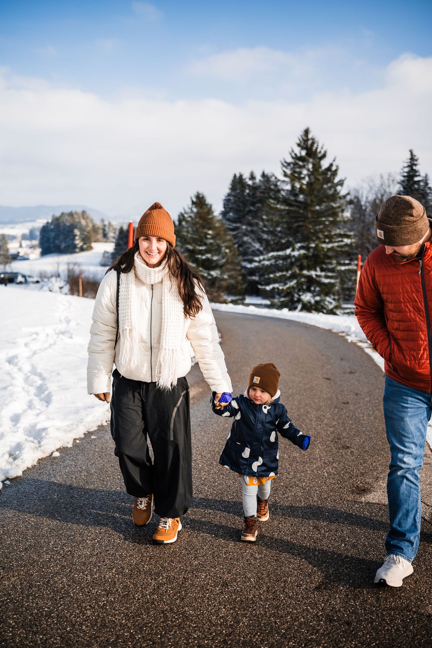 A family takes a winter walk together on a snowy path with evergreen trees in the background.