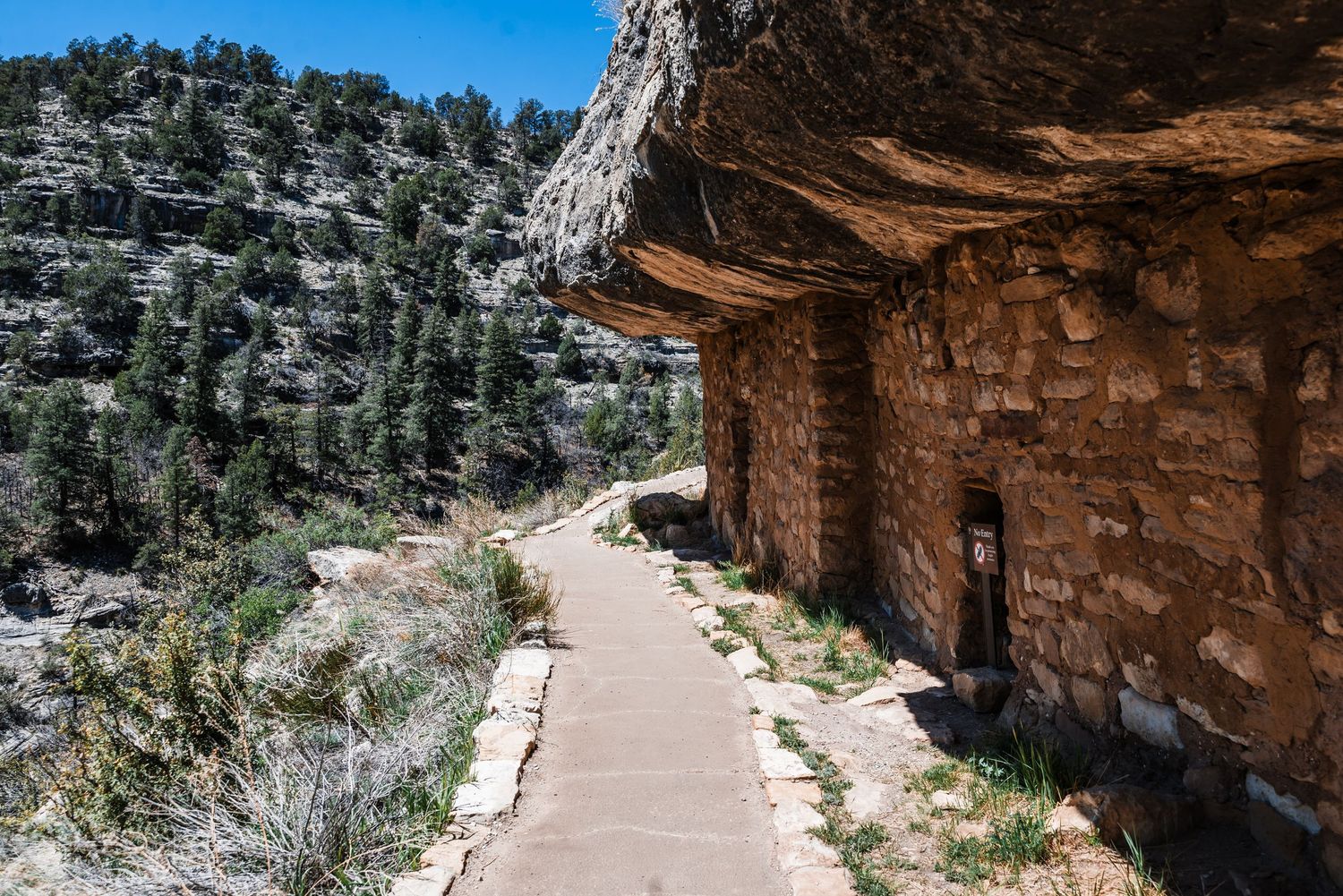 Paved trail winding along ancient cliff dwellings and rock formations in a sunny mountain landscape.