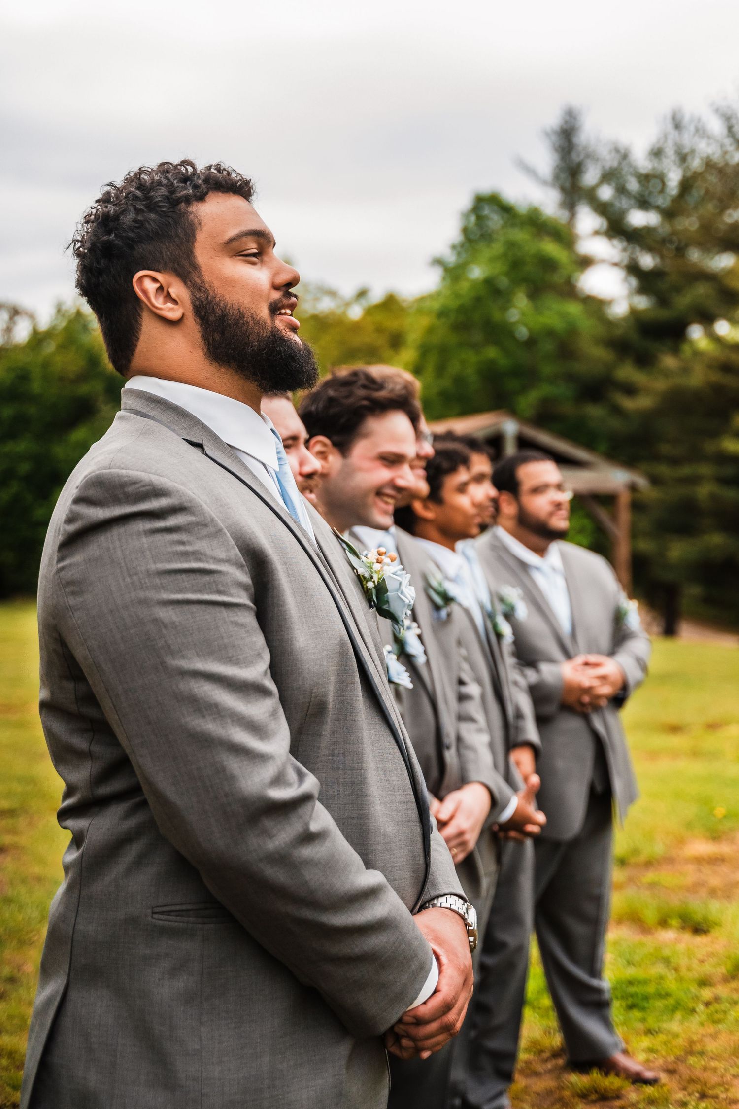 A smiling wedding party in grey suits stands outdoors against a backdrop of green trees and overcast sky.