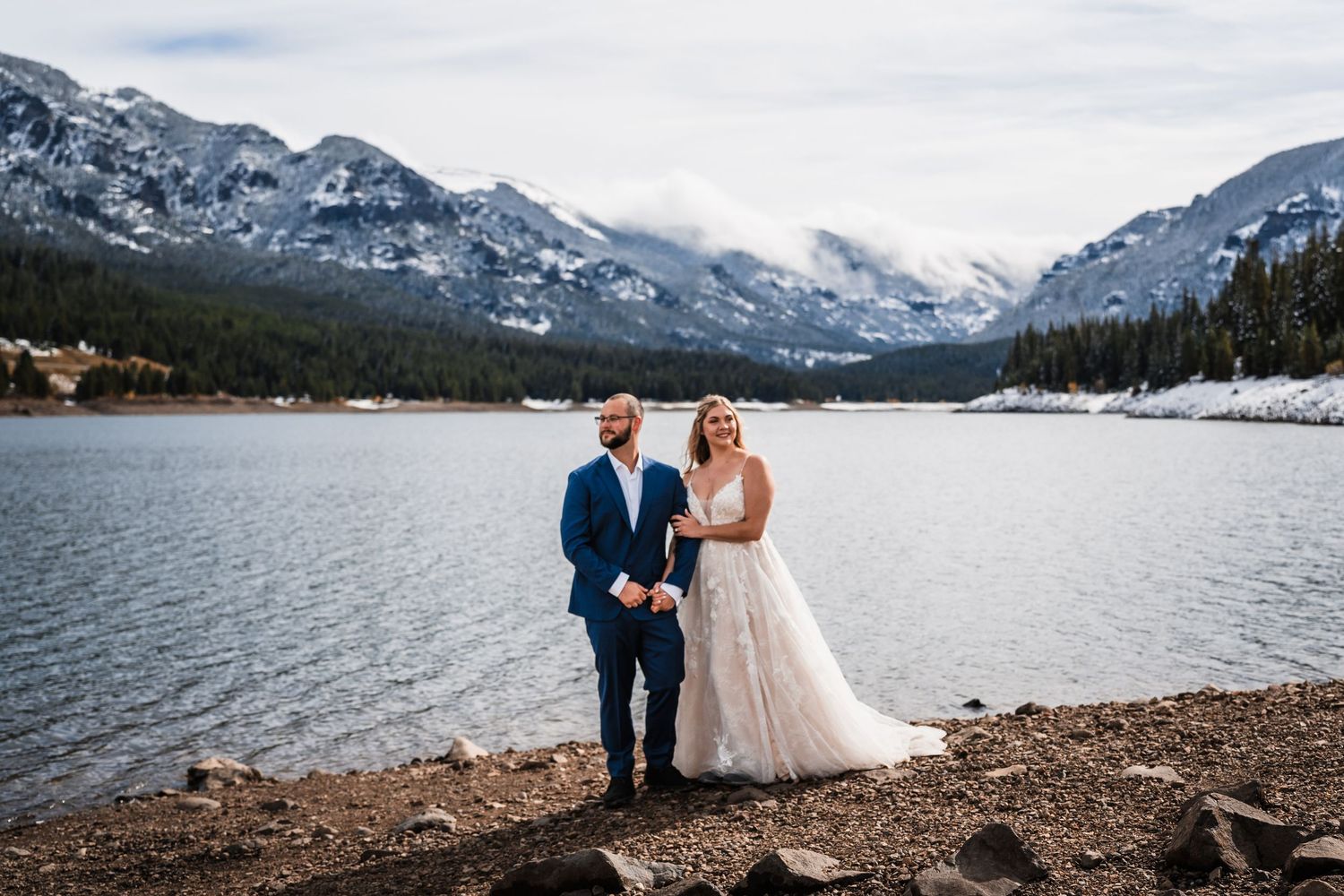 Wedding couple poses along mountain lake shore with snow-capped peaks in background.