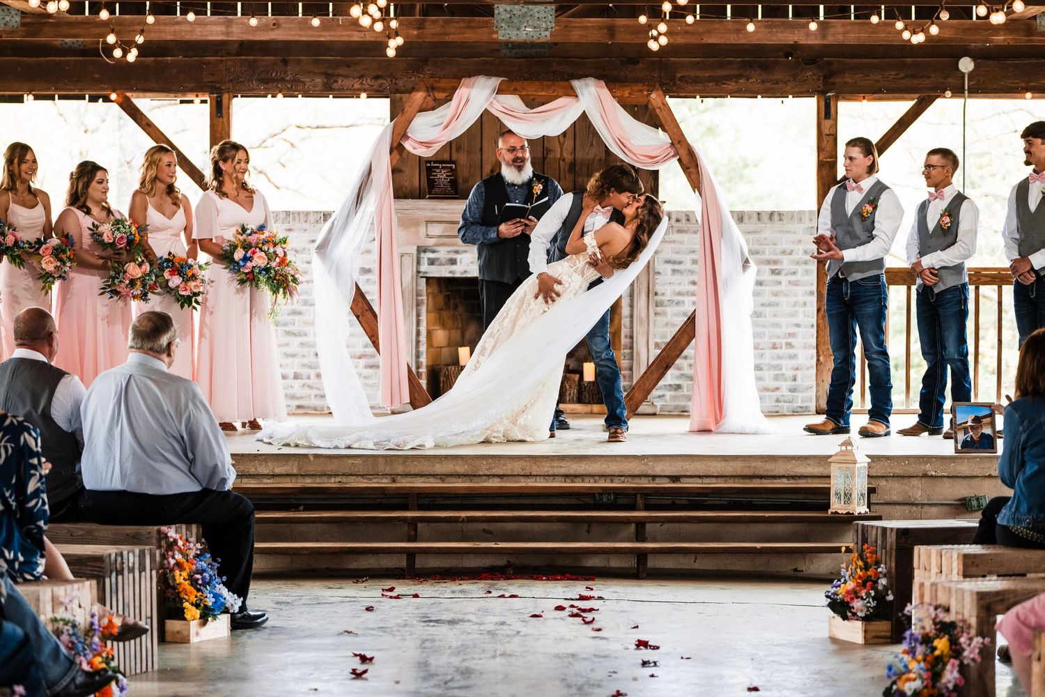 Celebratory moment during wedding ceremony under wooden beams with draped fabric and string lights.