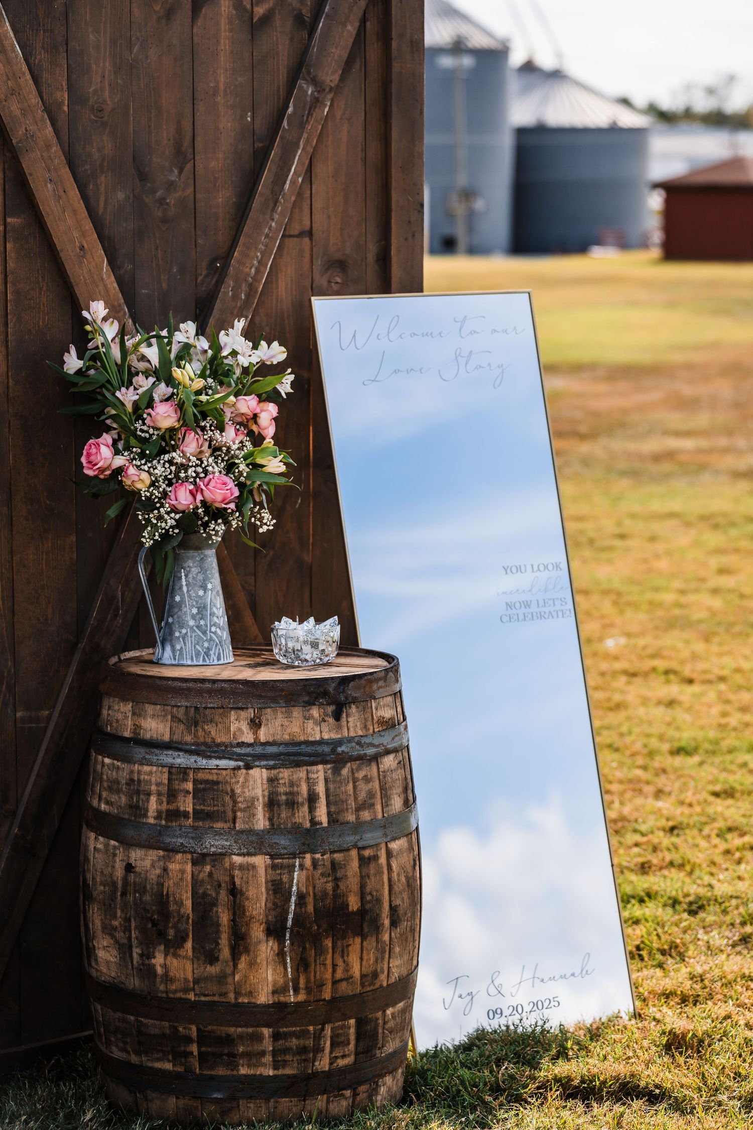 Rustic wedding decor featuring wooden barrels with floral arrangements and a mirror sign.