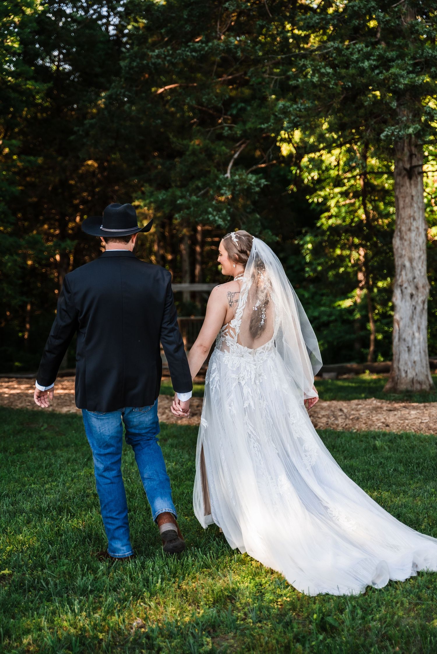 A couple walk through a wooded area, with the bride in white lace and her partner wearing a black shirt and jeans.