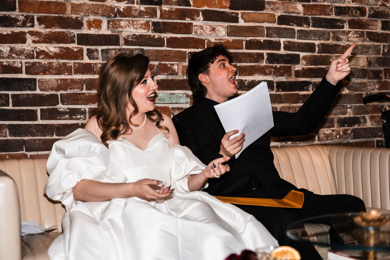 A couple seated on a leather couch against a brick wall share a joyful moment while looking at papers.