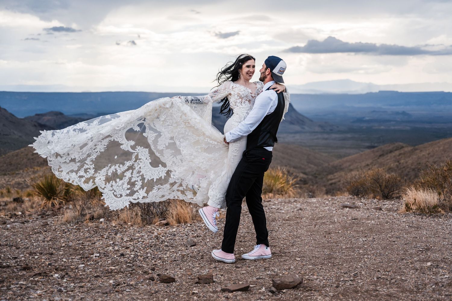 A couple embraces on a mountaintop as the bride's lace dress flows dramatically in the wind against mountain vistas.