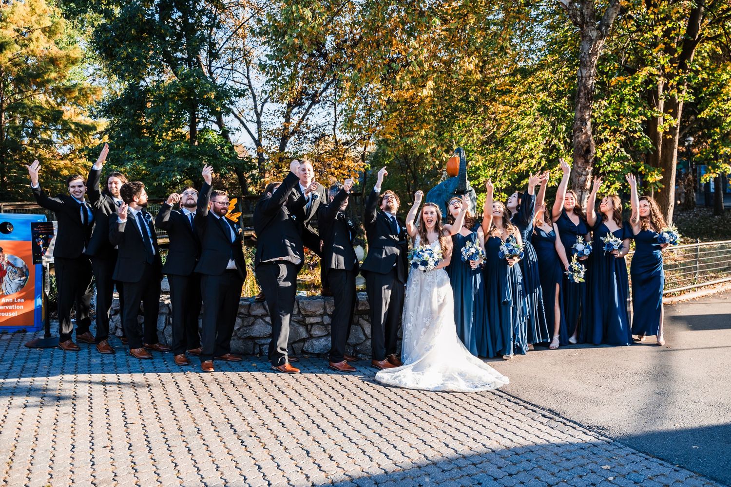 Large wedding party cheers and celebrates on brick pathway surrounded by fall foliage.