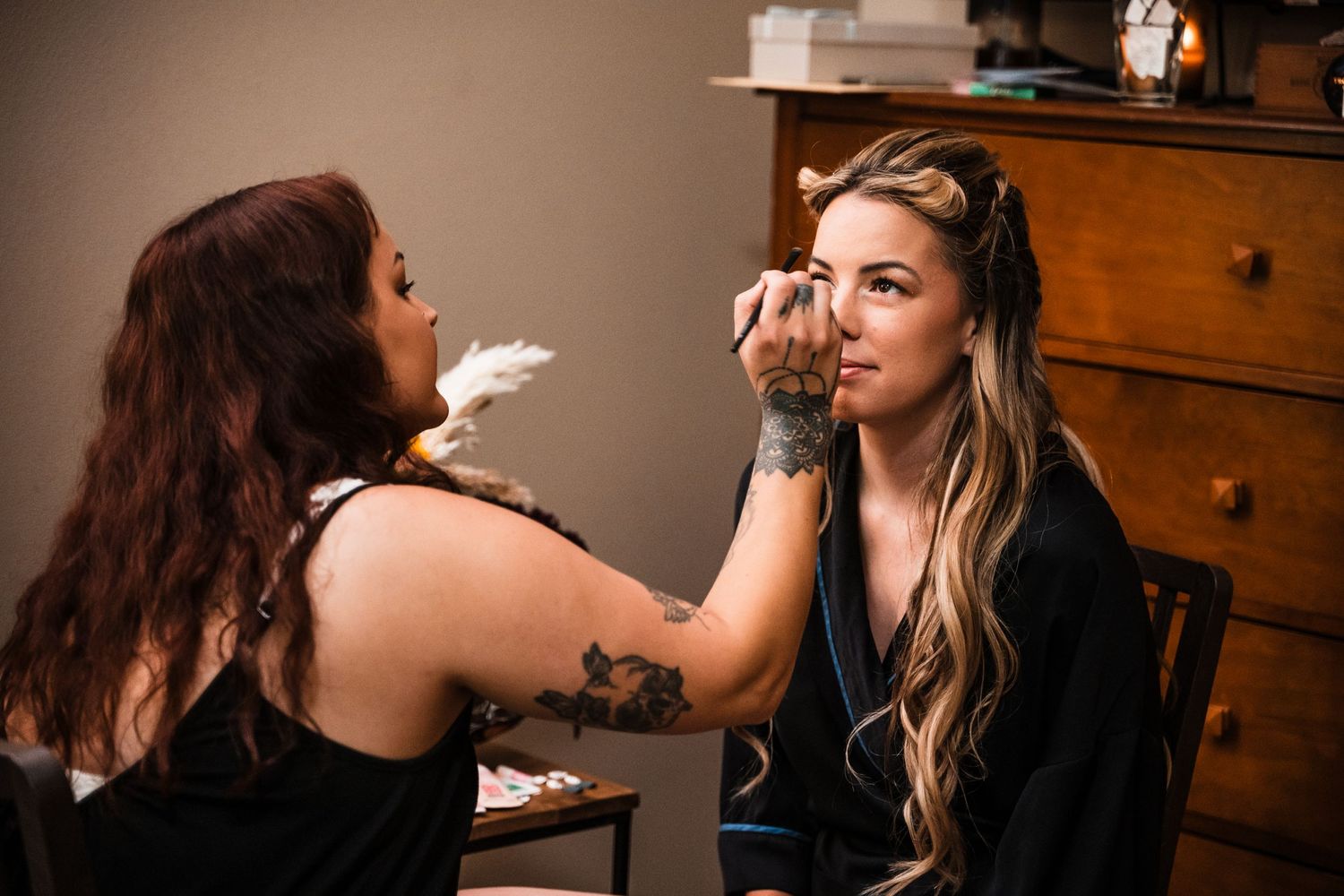 A makeup artist applies cosmetics to a client seated in a salon chair with wooden furniture in the background.