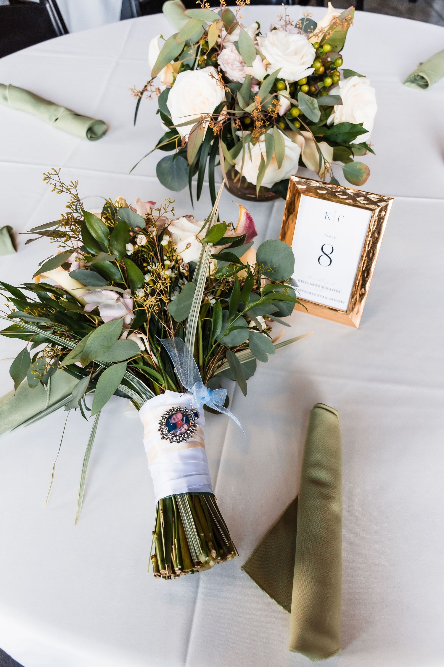 Elegant wedding table centerpiece with white roses and eucalyptus greenery next to a gold-framed table number.