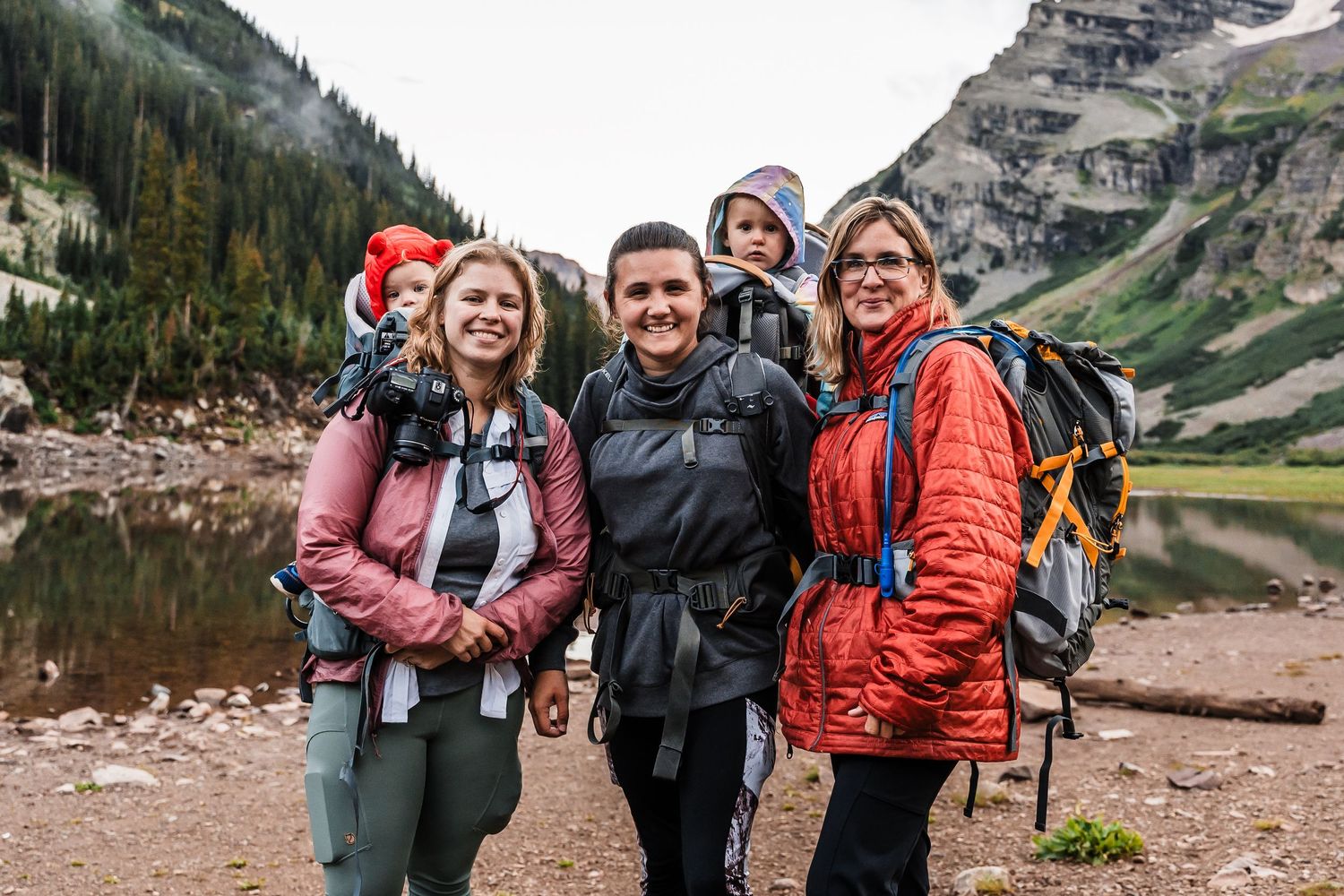 Group of hikers with backpacks standing near a mountain lake surrounded by rugged peaks and wilderness terrain.