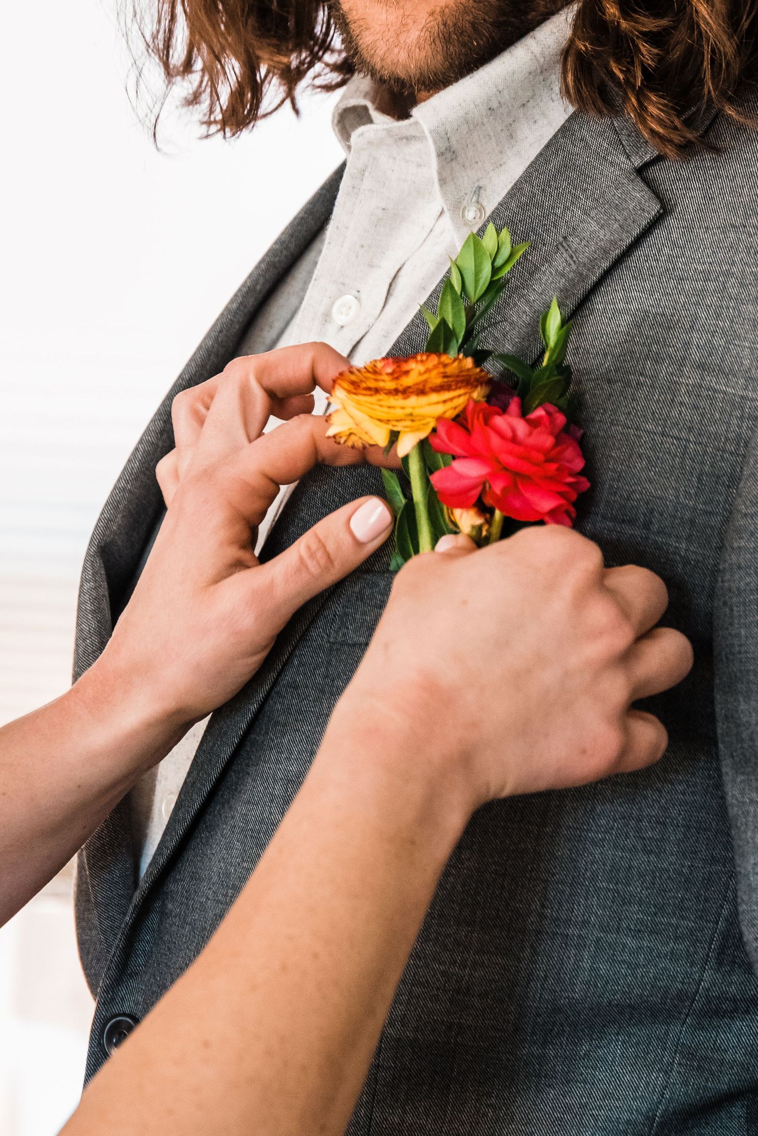 Hands pinning an orange and red floral boutonniere to a grey suit jacket.